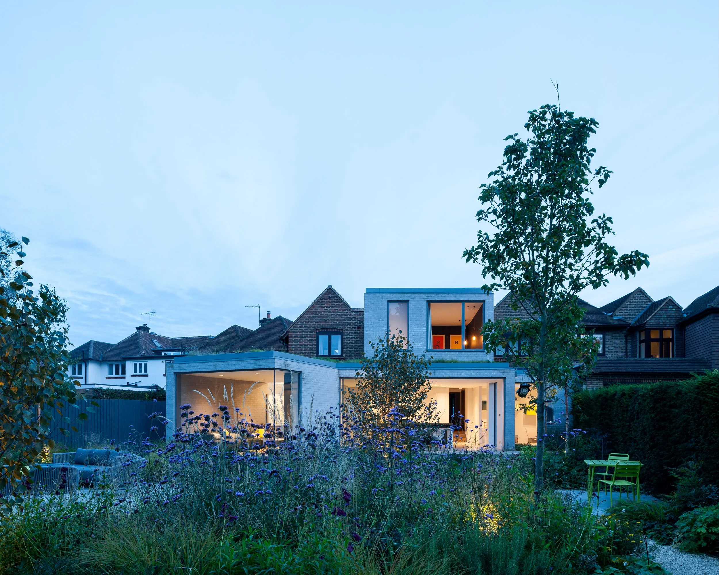 Modern multi-story house with large windows, illuminated from within, surrounded by a garden with various plants, trees, and outdoor furniture, during dusk.