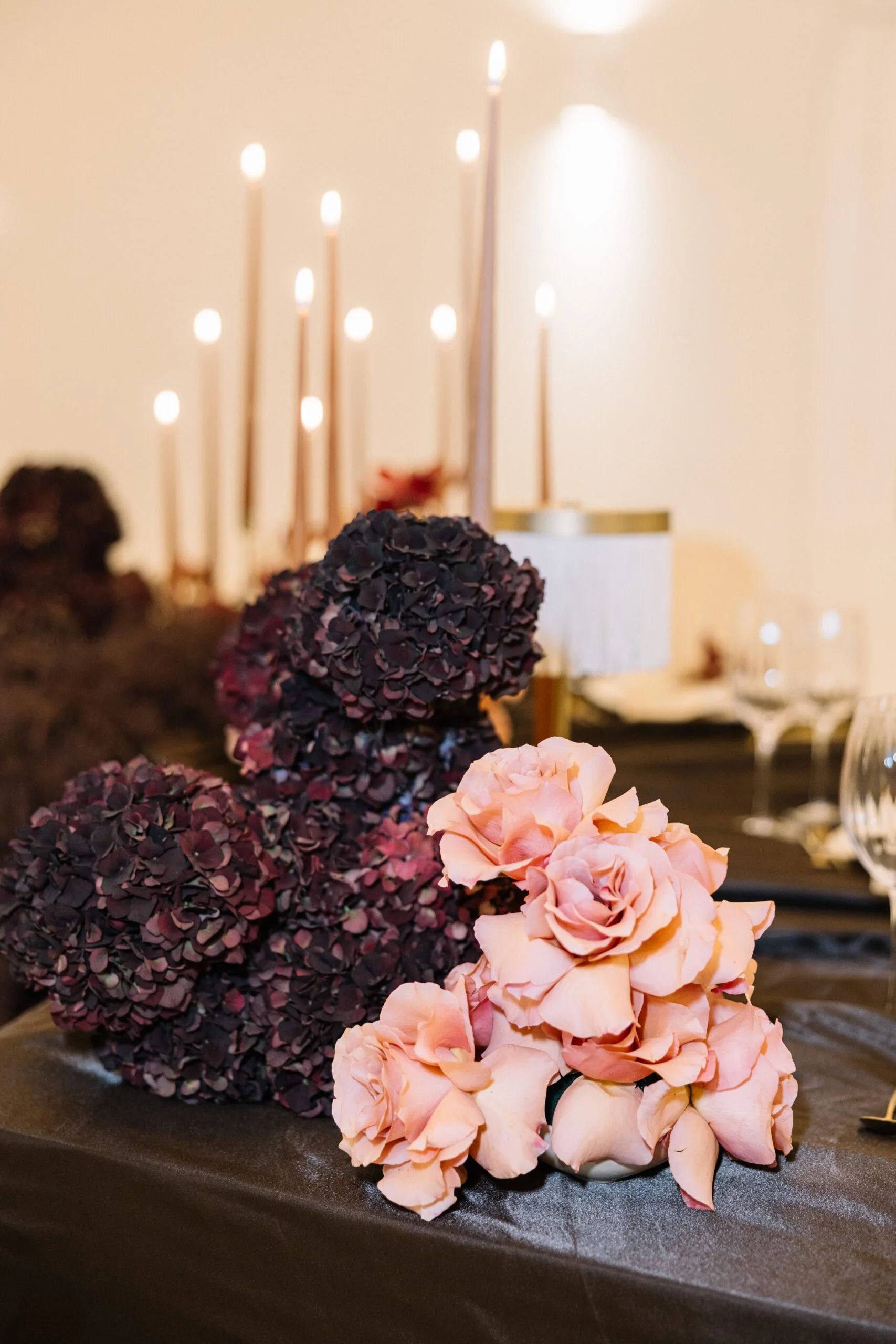A close-up of a floral centerpiece with pink roses and dark purple hydrangeas on a black tablecloth, with lit candles and glassware in the background.