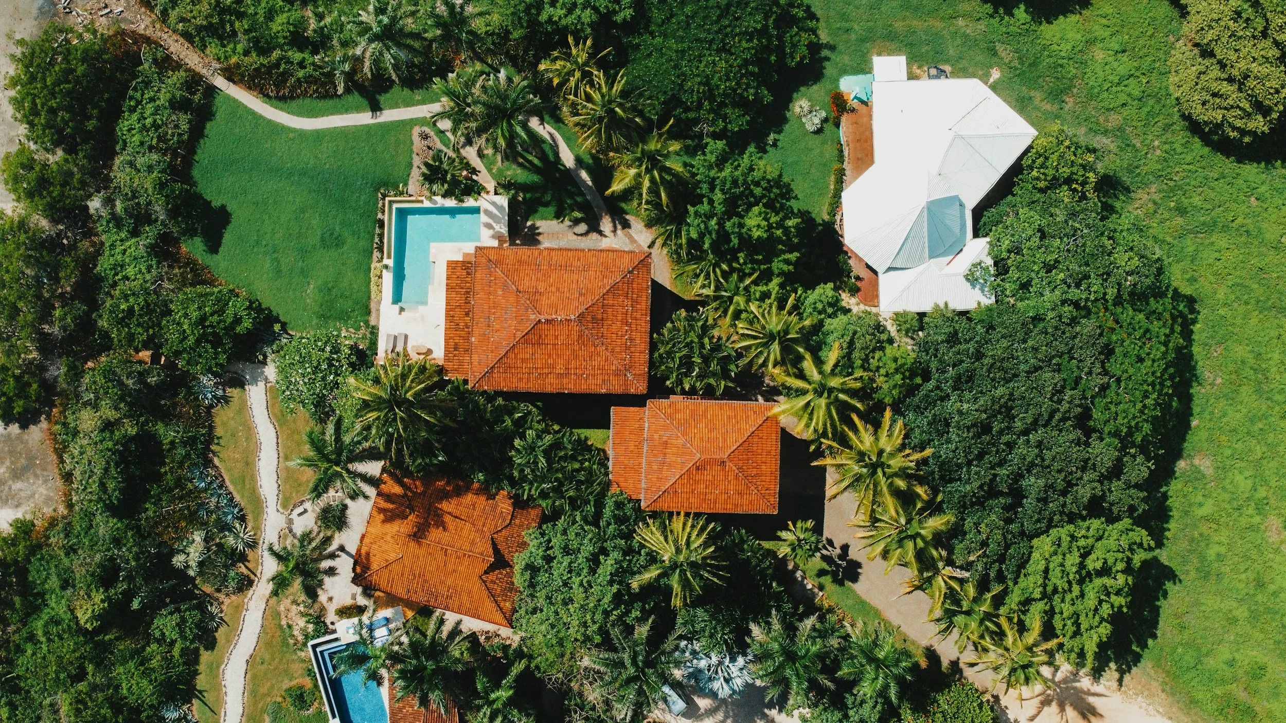 Aerial view of a tropical property with multiple houses, swimming pools, lush green trees, and lawns.