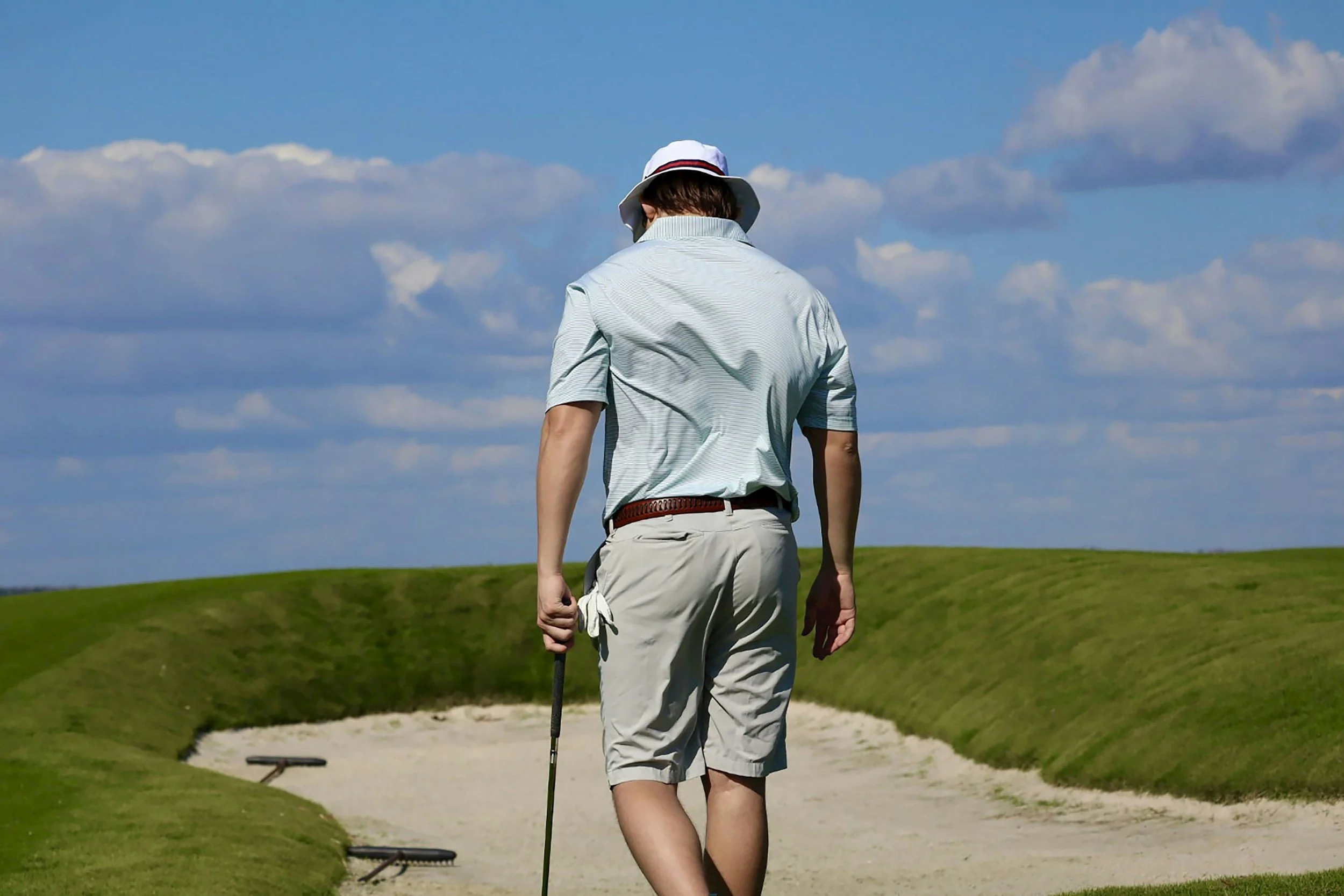 A man dressed in golf attire walking on a golf course sand trap, carrying a golf club, under a partly cloudy blue sky.