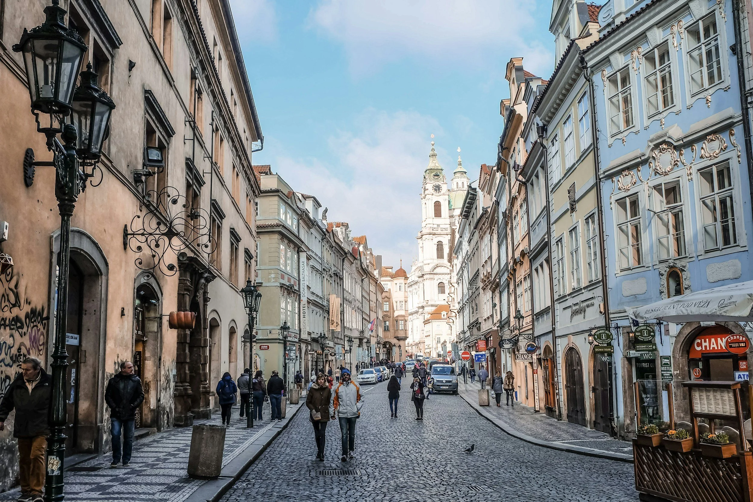 A cobblestone street in an old European city lined with historic pastel-colored buildings, some with graffiti, with pedestrians walking and cars parked, leading to a tall white church tower in the background on a partly cloudy day.