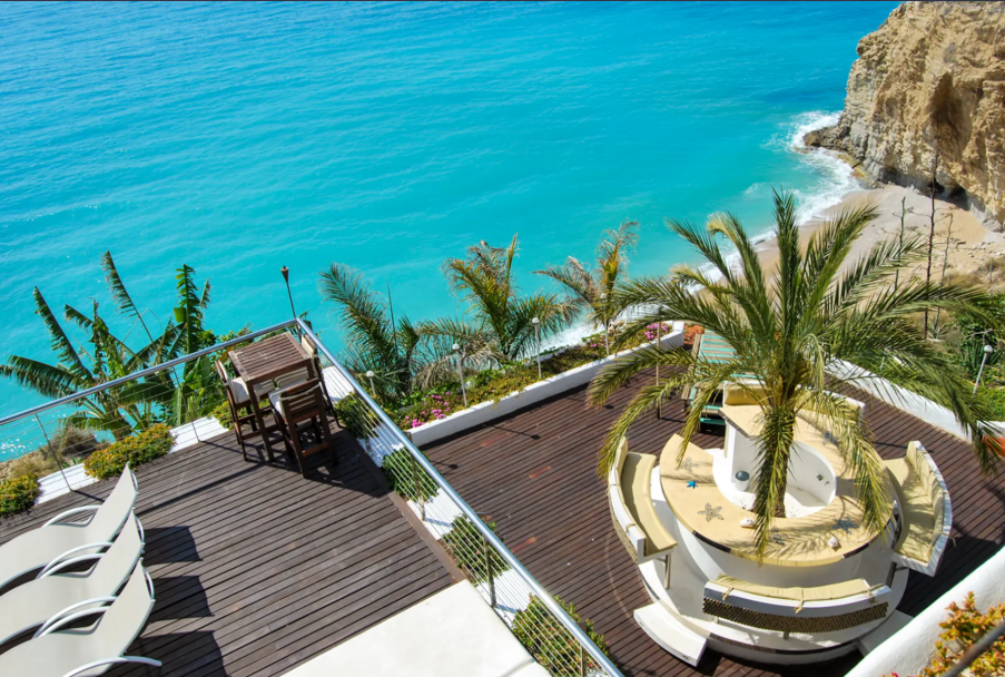 A rooftop terrace overlooking the ocean, decorated with palm trees, outdoor seating, and a wooden deck.
