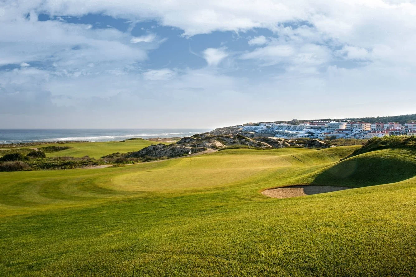 Scenic golf course near the coastline with green fairways, sand trap, and white buildings in the background under a partly cloudy sky.
