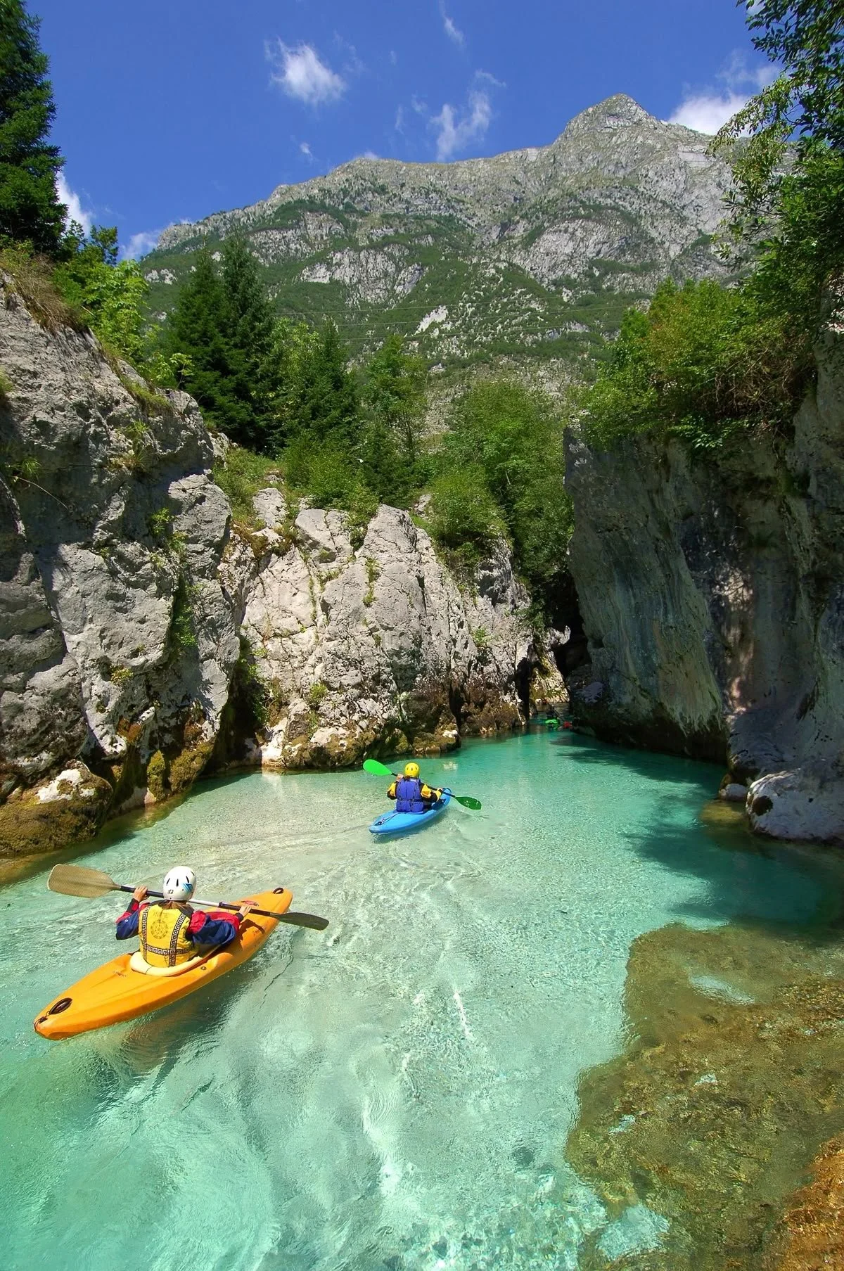 Two kayakers paddling through a narrow turquoise river canyon surrounded by rocky cliffs and green trees, with a mountain in the background under a partly cloudy blue sky.