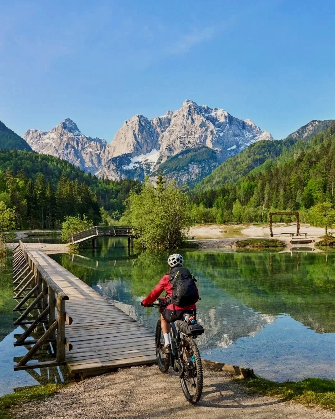 A person riding a mountain bike on a dirt path near a wooden bridge over a lake, with forested mountains and snow-capped peaks in the background.