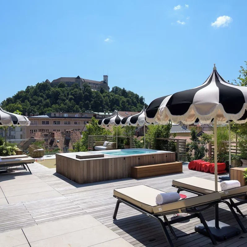 Rooftop pool with lounge chairs and black-and-white striped umbrellas, overlooking a hillside cityscape with a castle on a hill in the background.