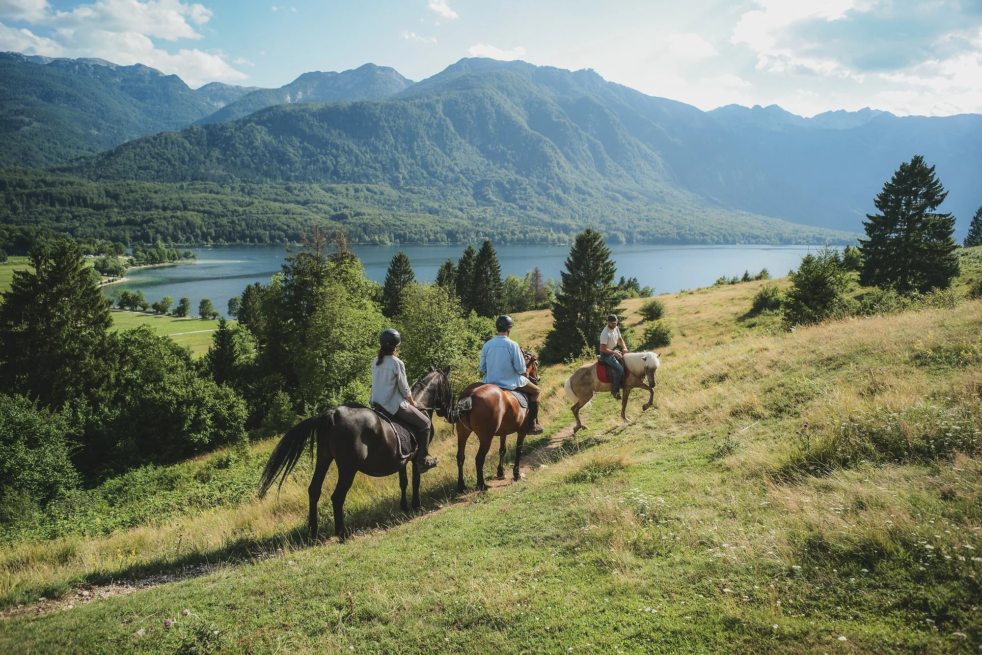 Four people riding horses along a grassy trail with a lake and mountains in the background under partly cloudy skies.