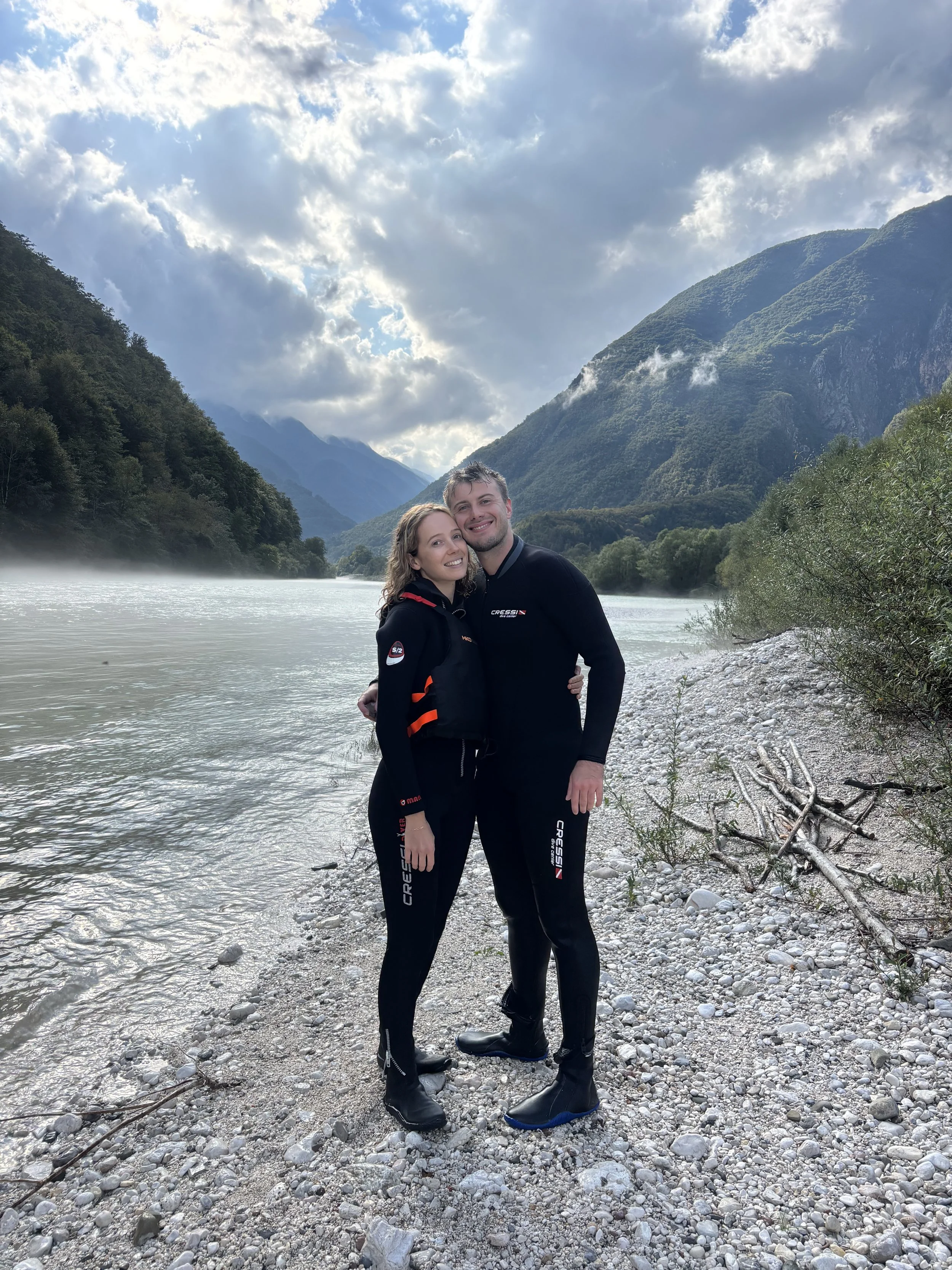 A couple wearing black Cressi wetsuits standing on a pebbled riverbank with mountainous terrain and cloudy sky in the background.