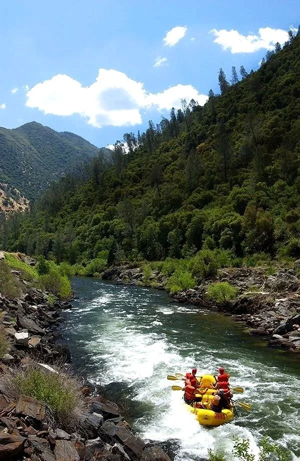 Four people in a yellow inflatable raft with red life jackets and paddles rafting on a river surrounded by lush green mountains and a partly cloudy sky.