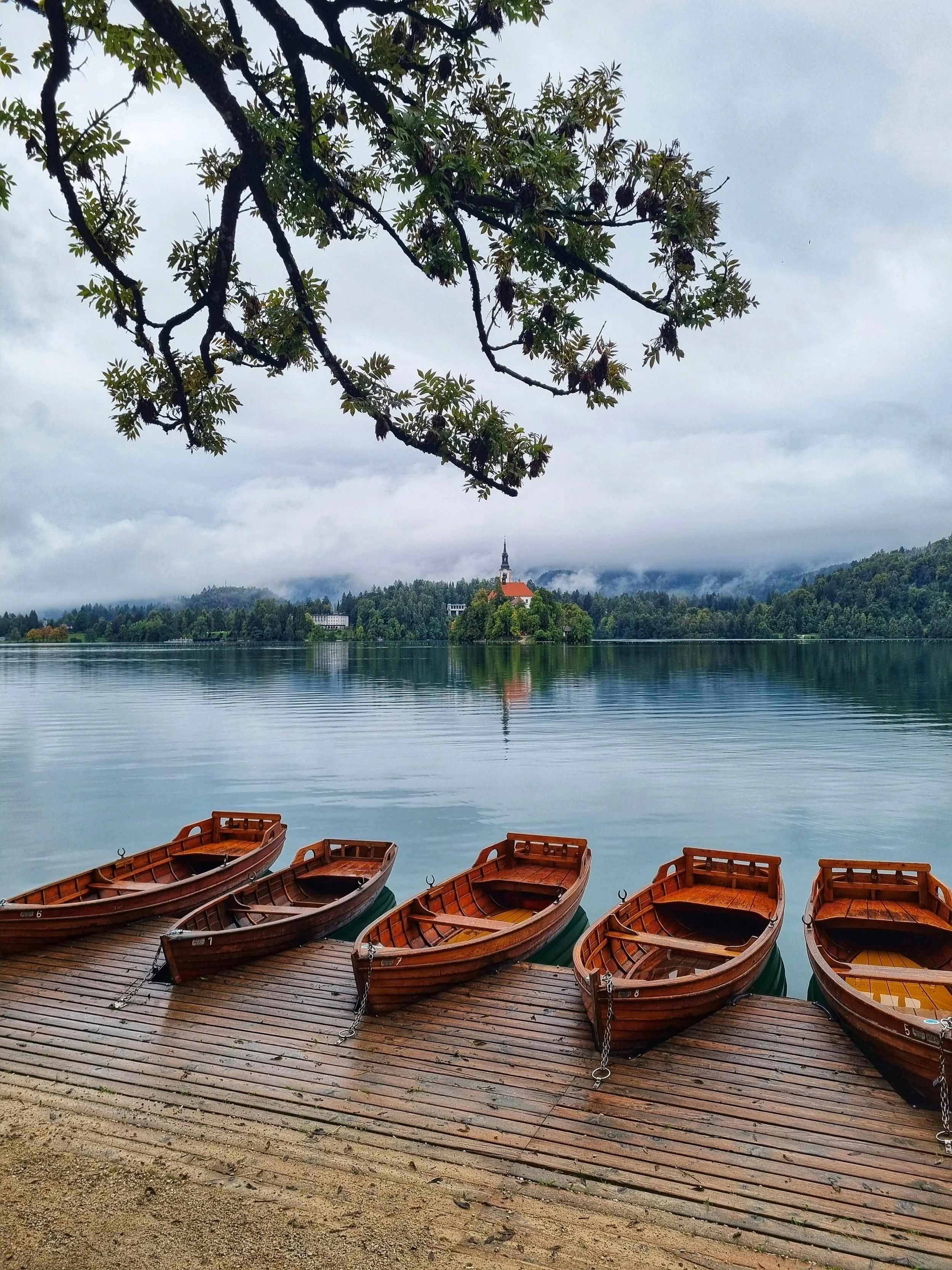 Five wooden rowboats tied to a dock on a calm lake, with a church on an island and forested hills in the background under cloudy sky.