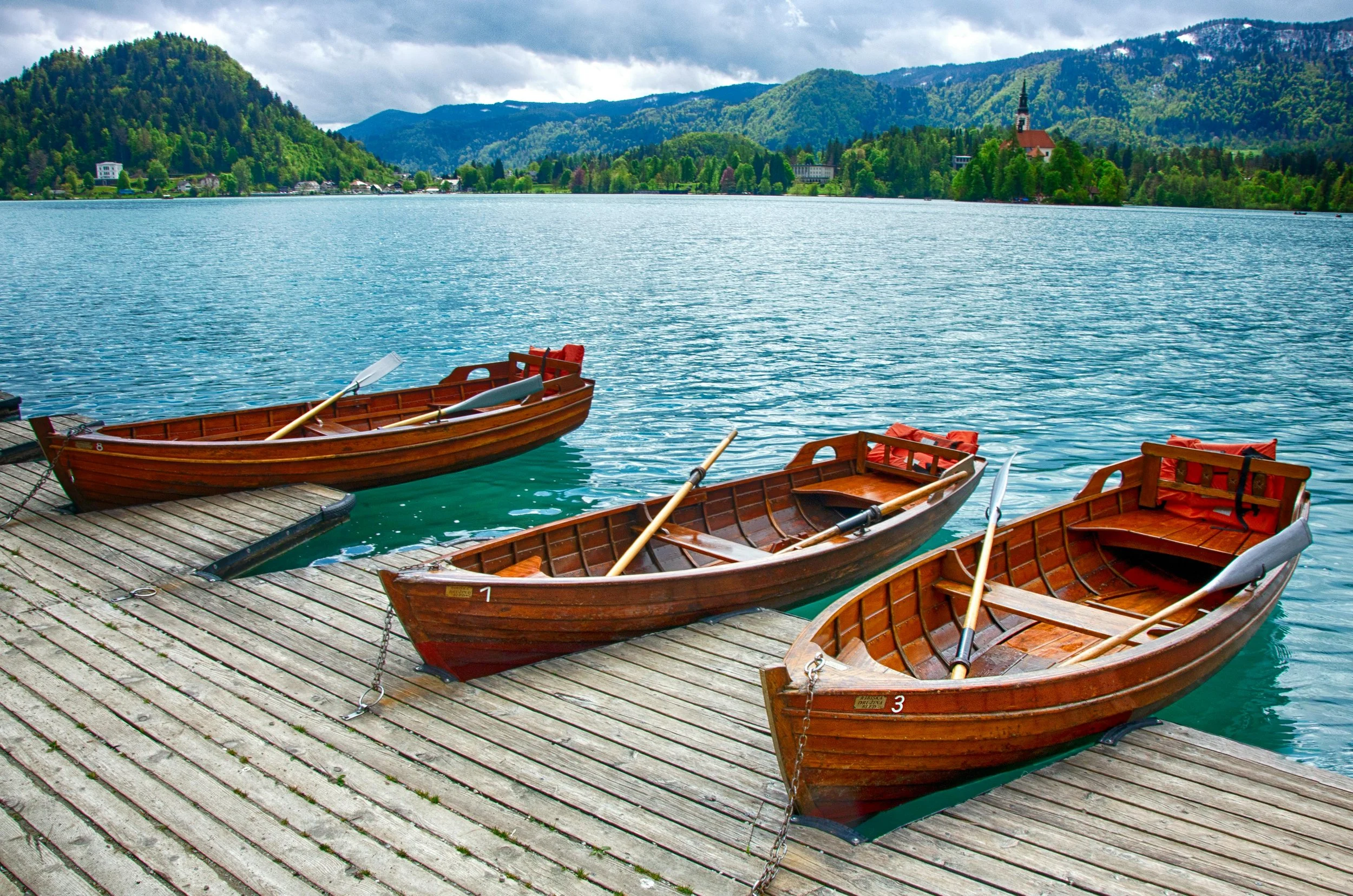 Three wooden rowboats tied to a dock on a lake, with mountains and a church in the background.
