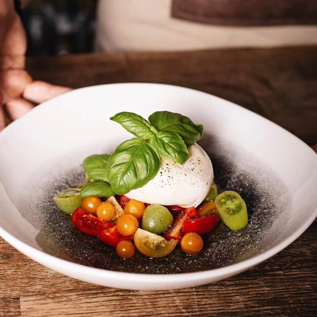Caprese salad with cherry tomatoes, sliced green tomatoes, basil leaves, mozzarella ball, black pepper, and balsamic vinegar on a white plate.