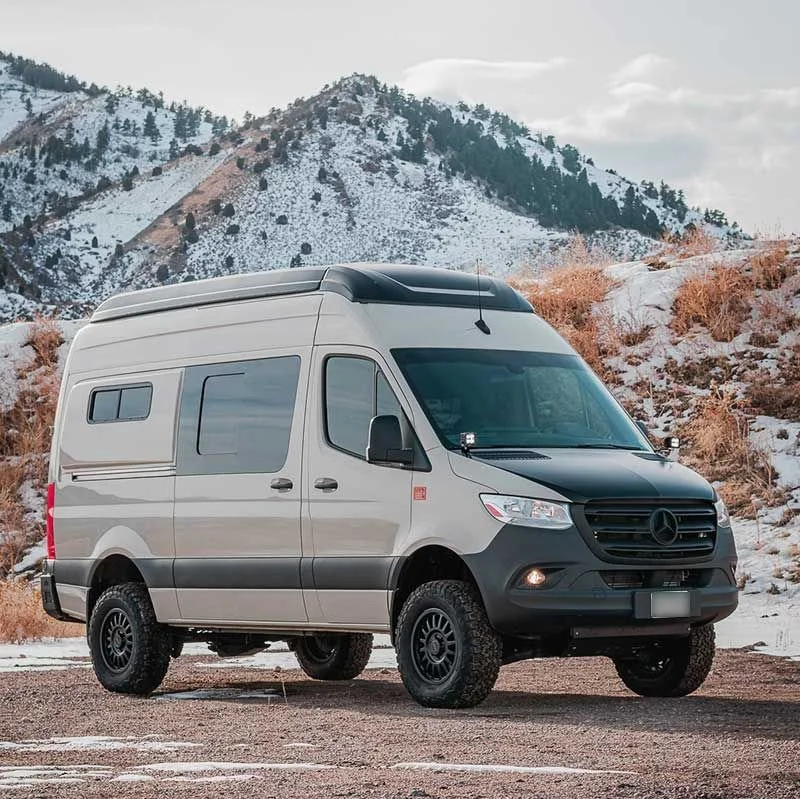 A gray Mercedes-Benz camper van on a gravel lot with snow-covered mountains in the background.