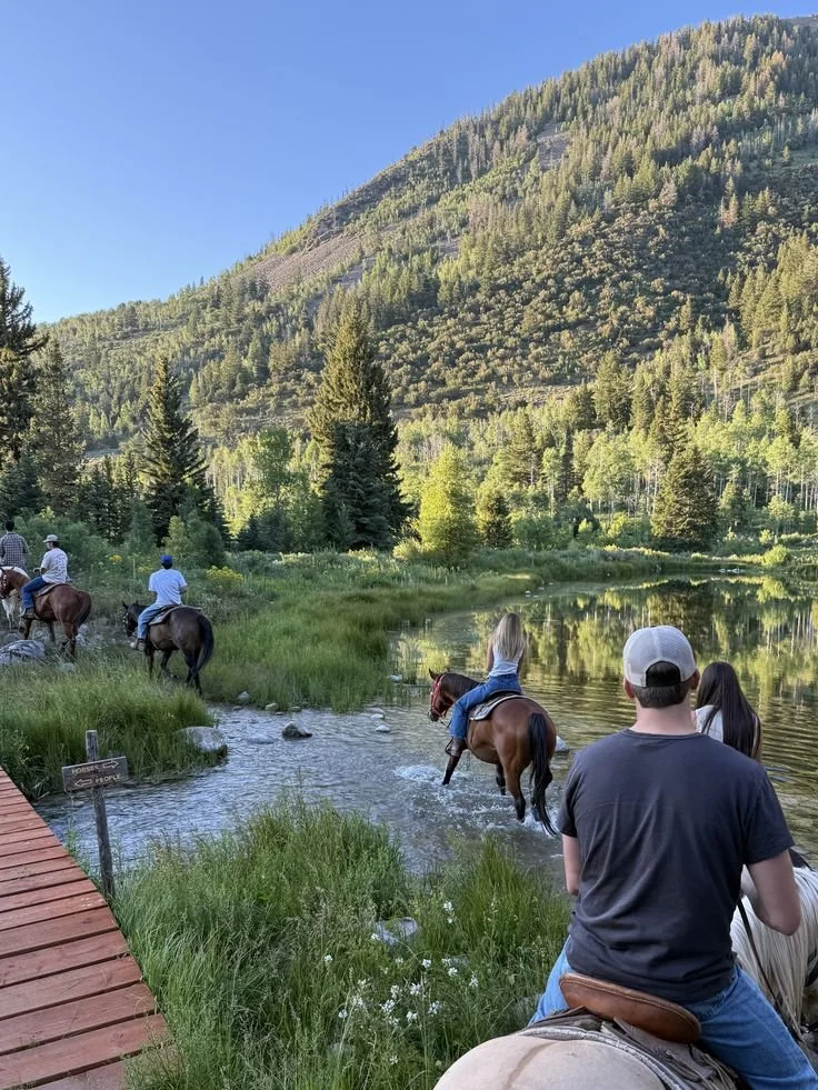 People horseback riding near a river in a lush, green mountainous landscape under a clear blue sky.