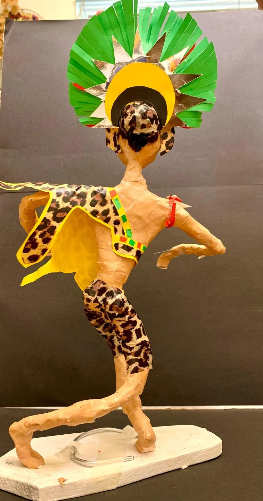 A colorful paper-mâché sculpture of a person dressed as a traditional African tribal dancer, wearing a leopard-patterned outfit and a large feathered headdress with green, yellow, and silver accents.