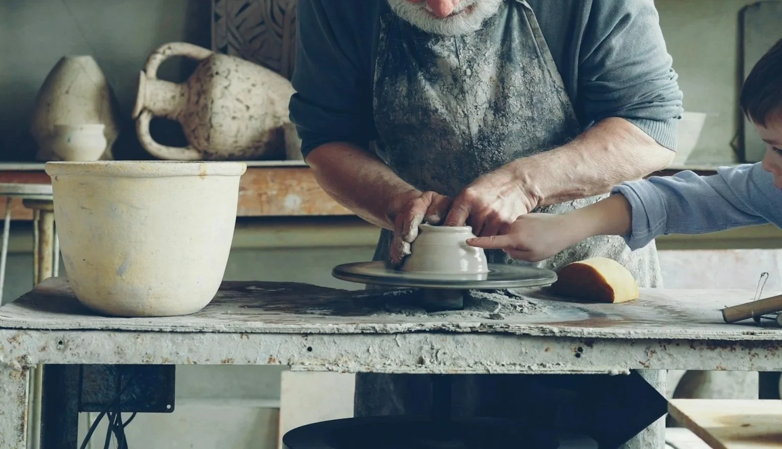 A person and a child are shaping a clay pot on a pottery wheel in a pottery studio. The person is wearing a dirty apron and a long-sleeve shirt, with their hands on the clay. The child's hand is touching the pot. There are various pottery tools and unfinished clay pieces on the work table.