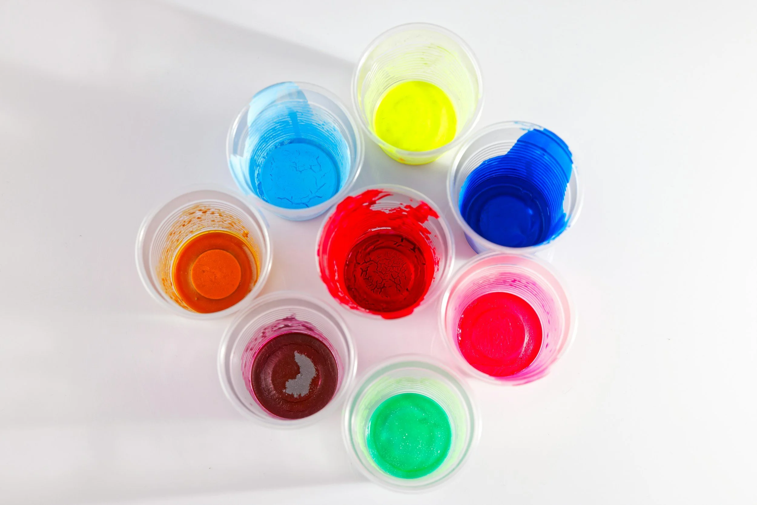 Top-down view of nine plastic cups filled with various colors of liquid, including yellow, blue, red, pink, orange, green, and purple, on a white surface.