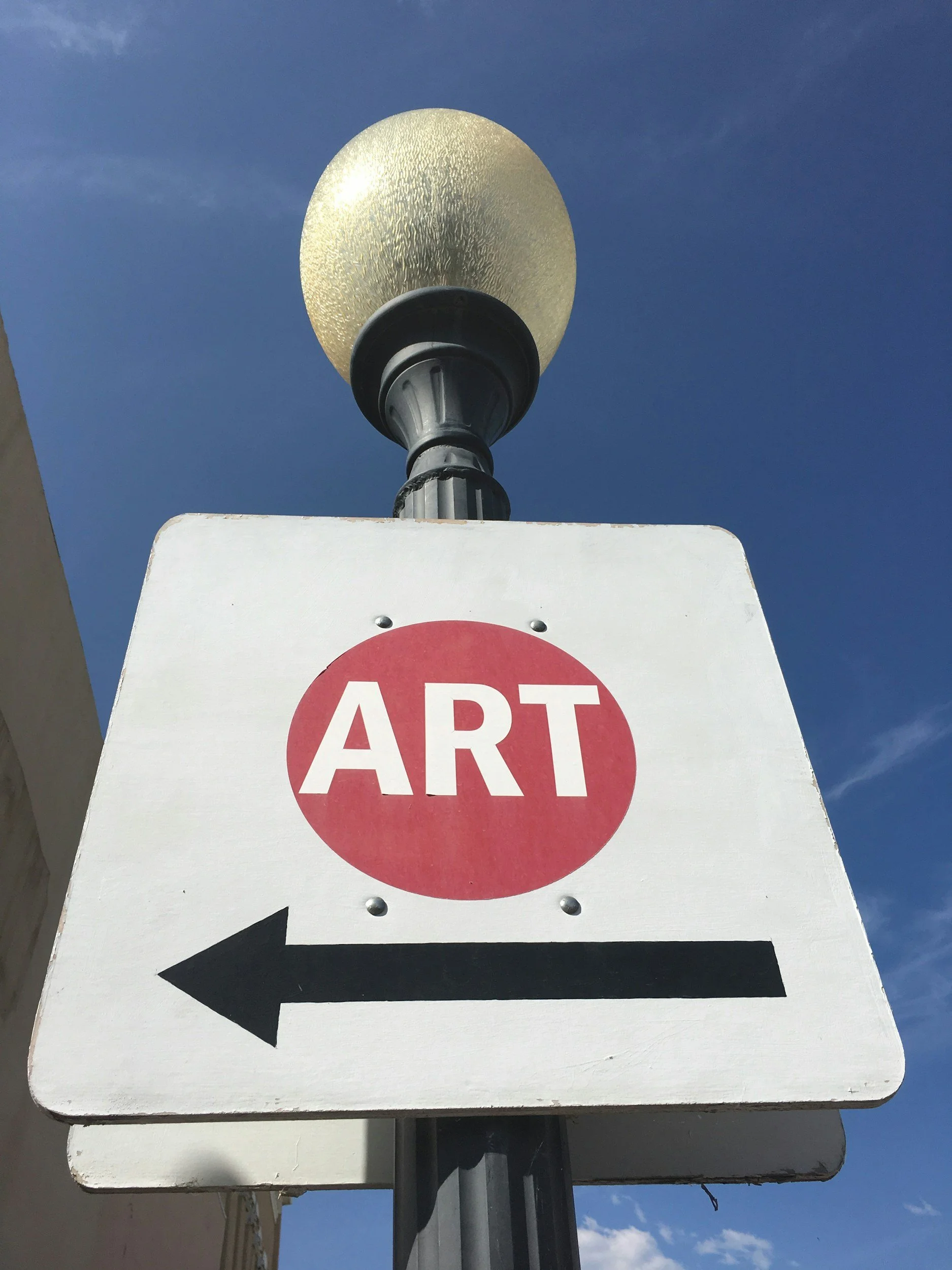 A street sign with the word 'ART' in a red circle and an arrow pointing left, mounted on a pole beneath a textured globe-shaped lamp, against a blue sky.