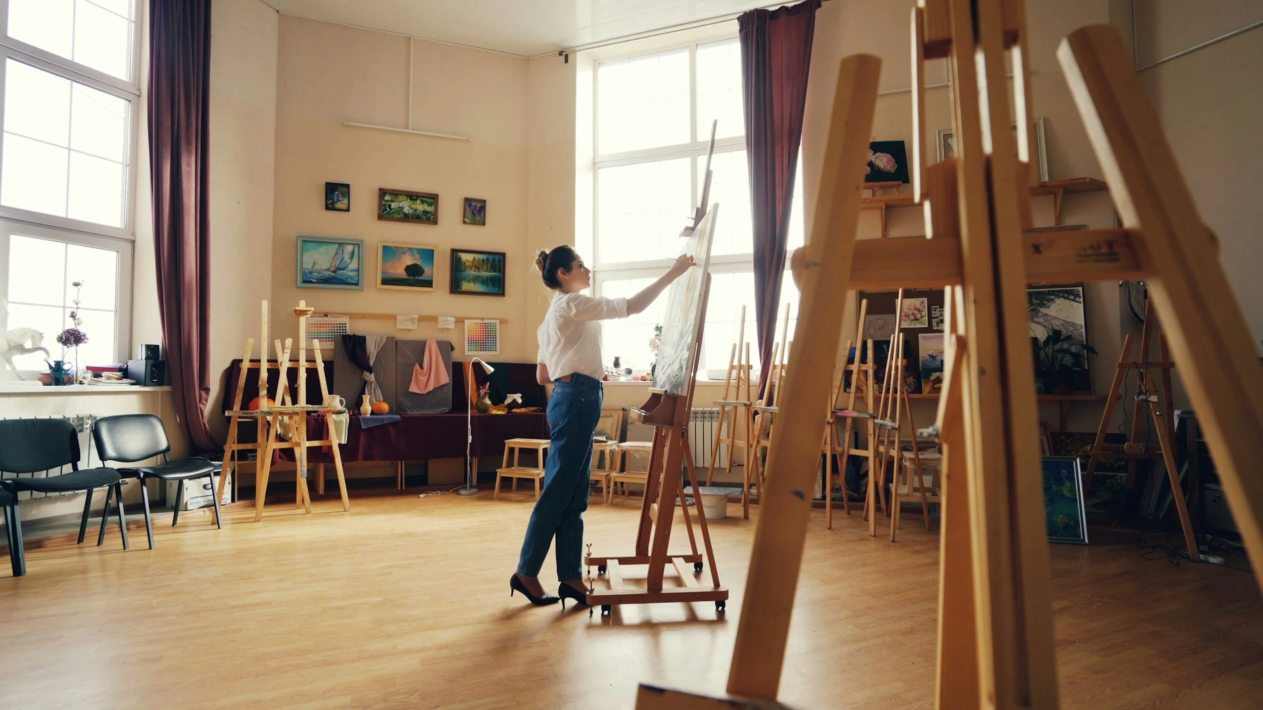 A woman painting on a large canvas in an art studio filled with easels, paintings, and art supplies.