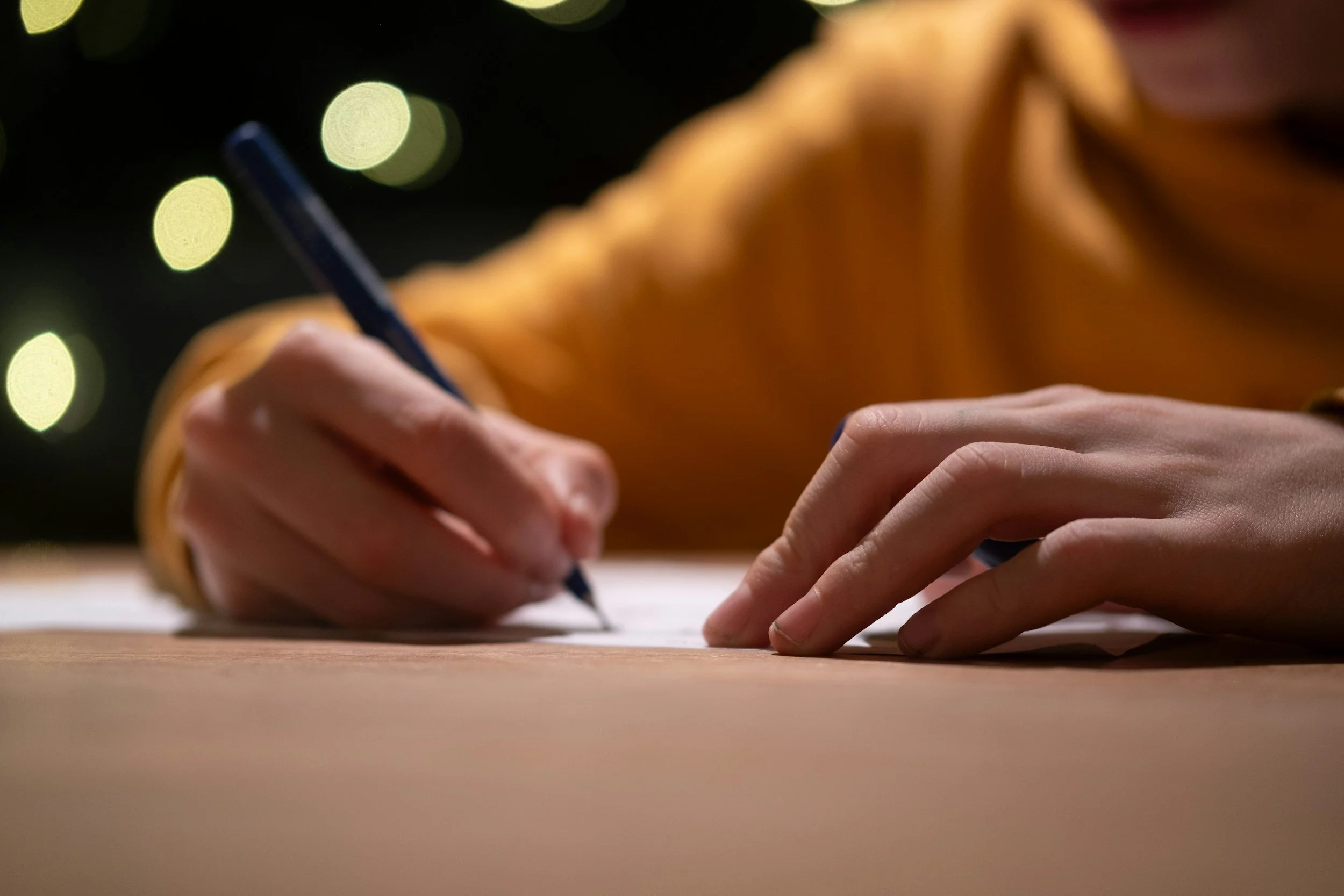 A person wearing an orange sweater is writing on a piece of paper with a dark pen, with a blurred background of circular yellow lights.