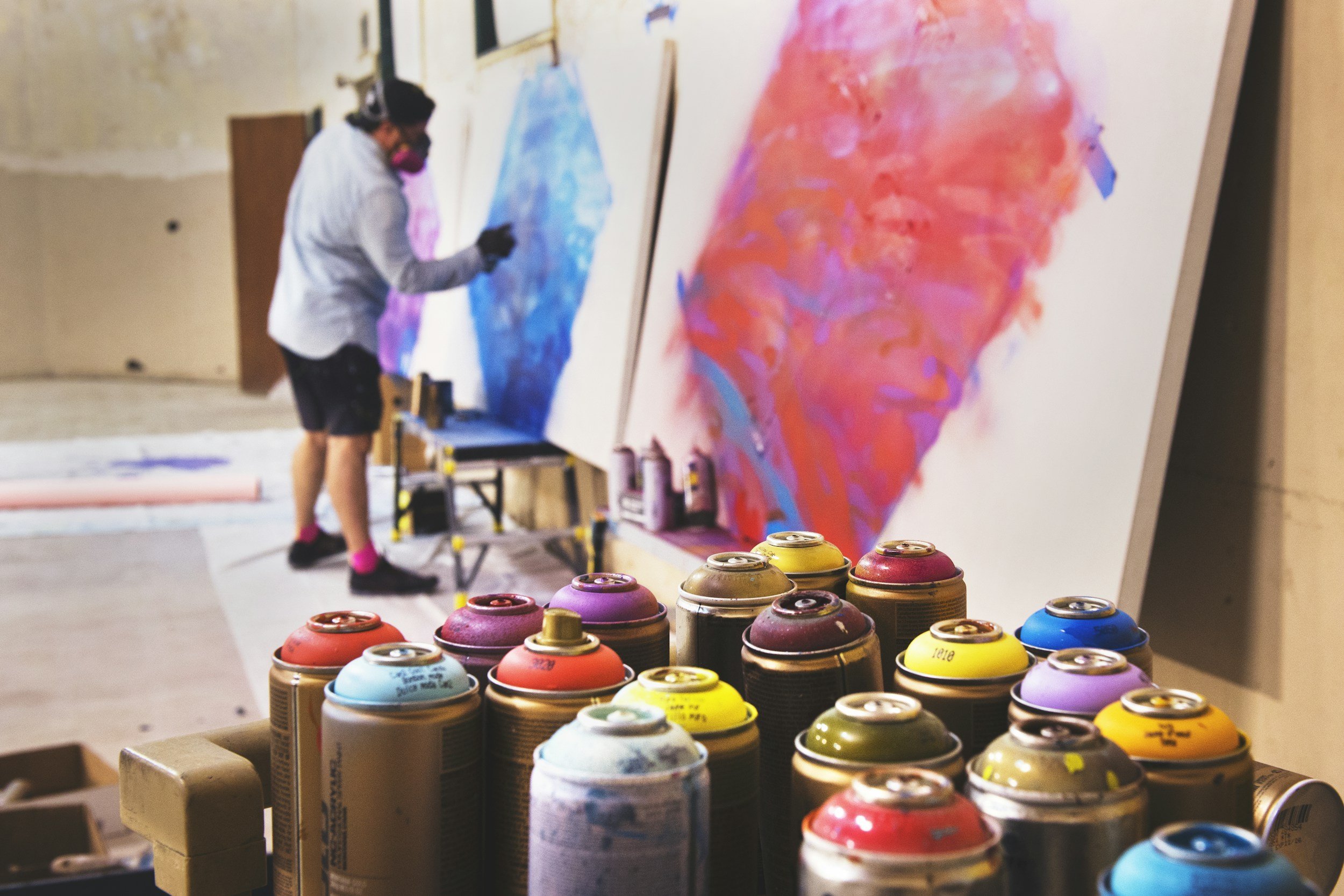 An artist is spray painting a large canvas with shades of purple and pink inside a studio. Multiple spray paint cans are arranged in the foreground.