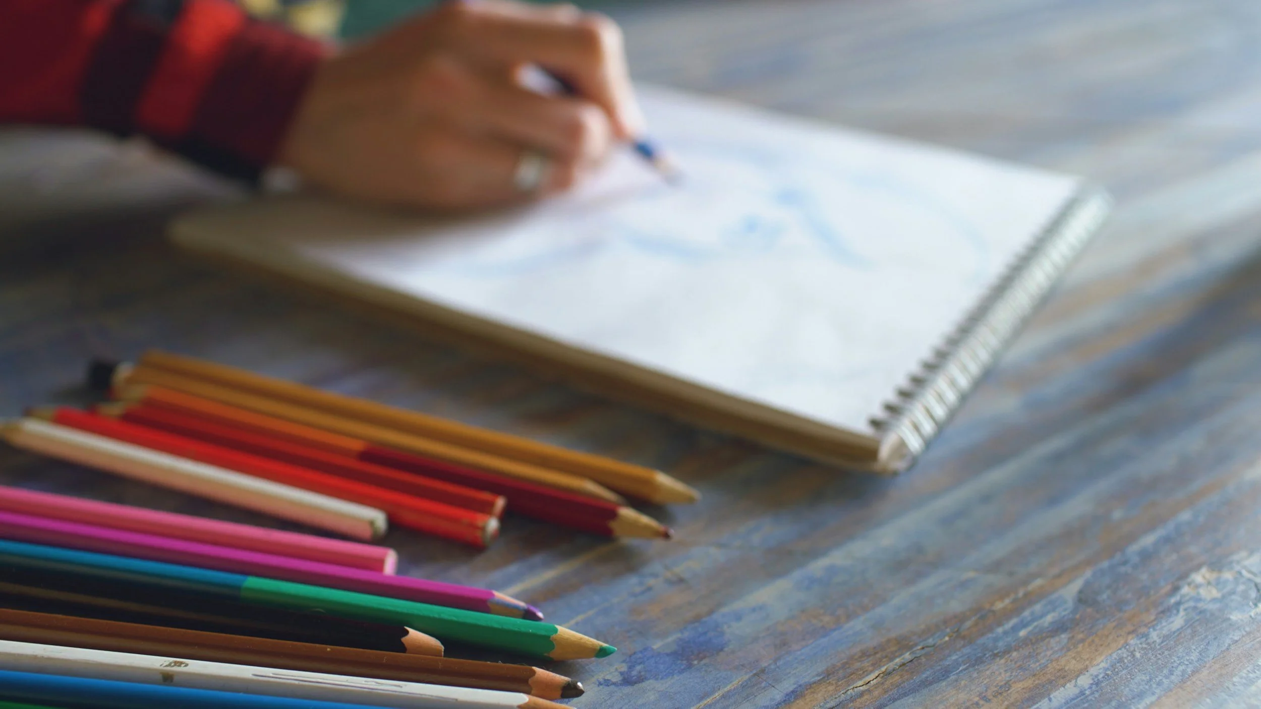 A person's hand drawing in a spiral notebook with colored pencils arranged on a wooden table.