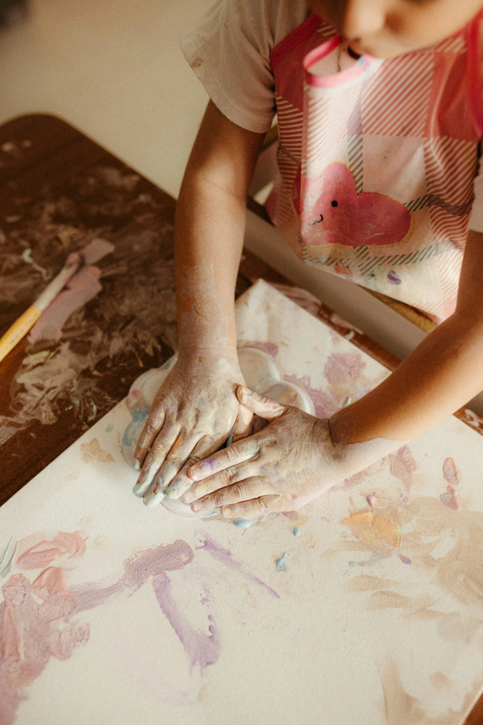Child shaping wet clay on a table, hands covered in clay, using a small sculpting tool, wearing a pink apron with a flamingo design.