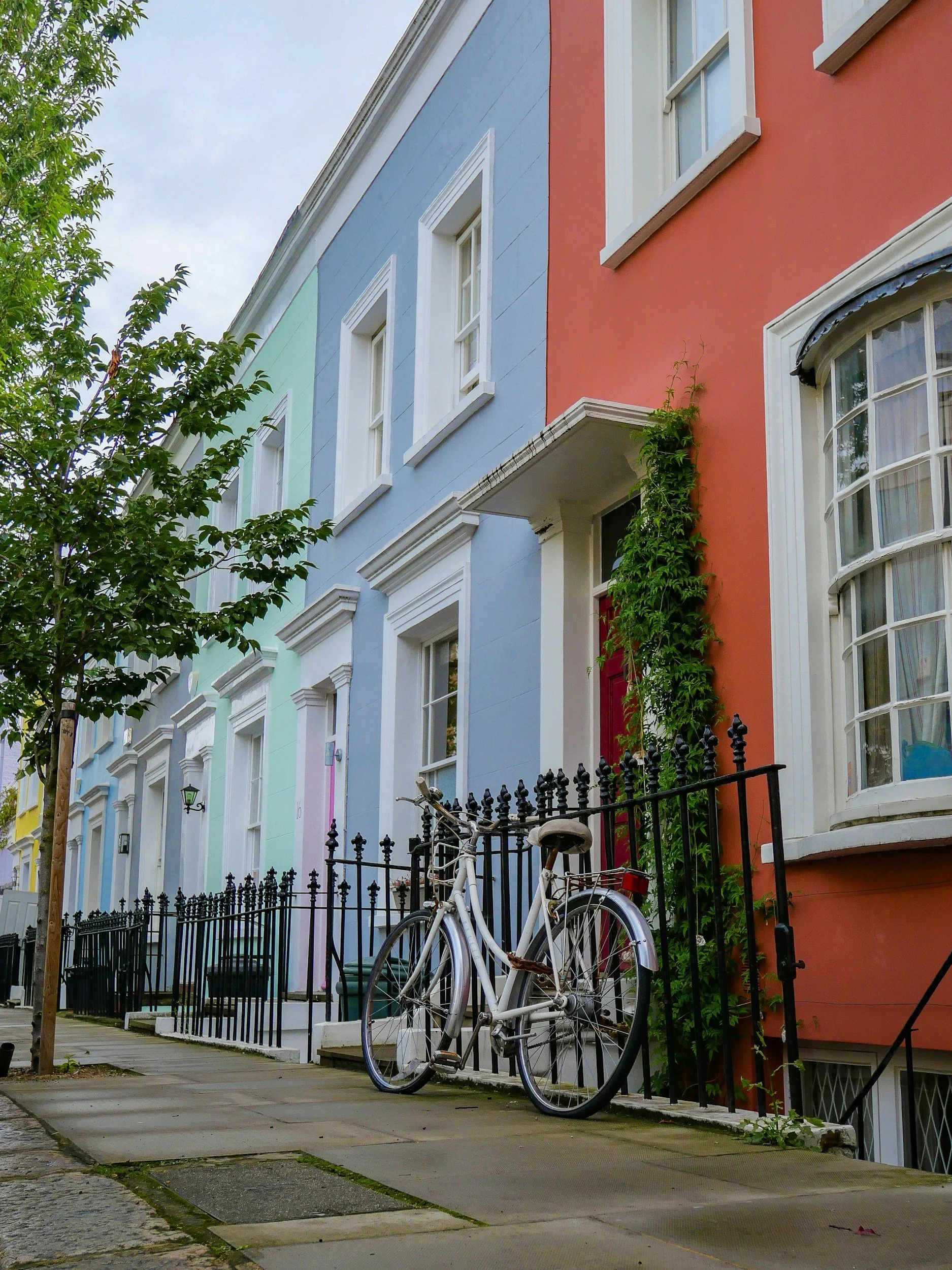 Colorful row houses with pastel facades on a city street, a white bicycle parked against a black fence, a tree on the sidewalk, and a cloudy sky.