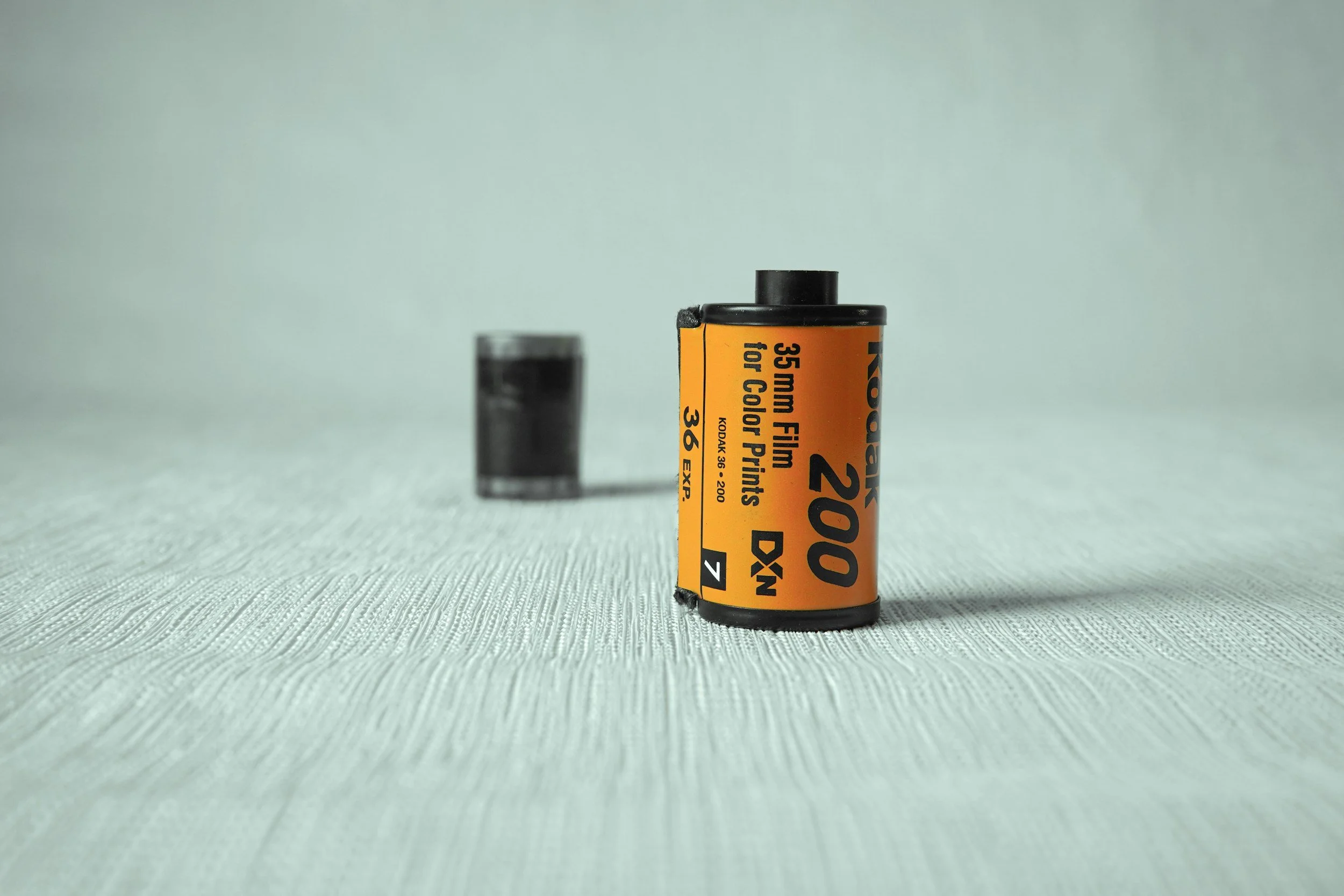 A roll of Kodak 200 film with an orange label and black text, standing upright on a textured white surface with a blurred black can in the background.