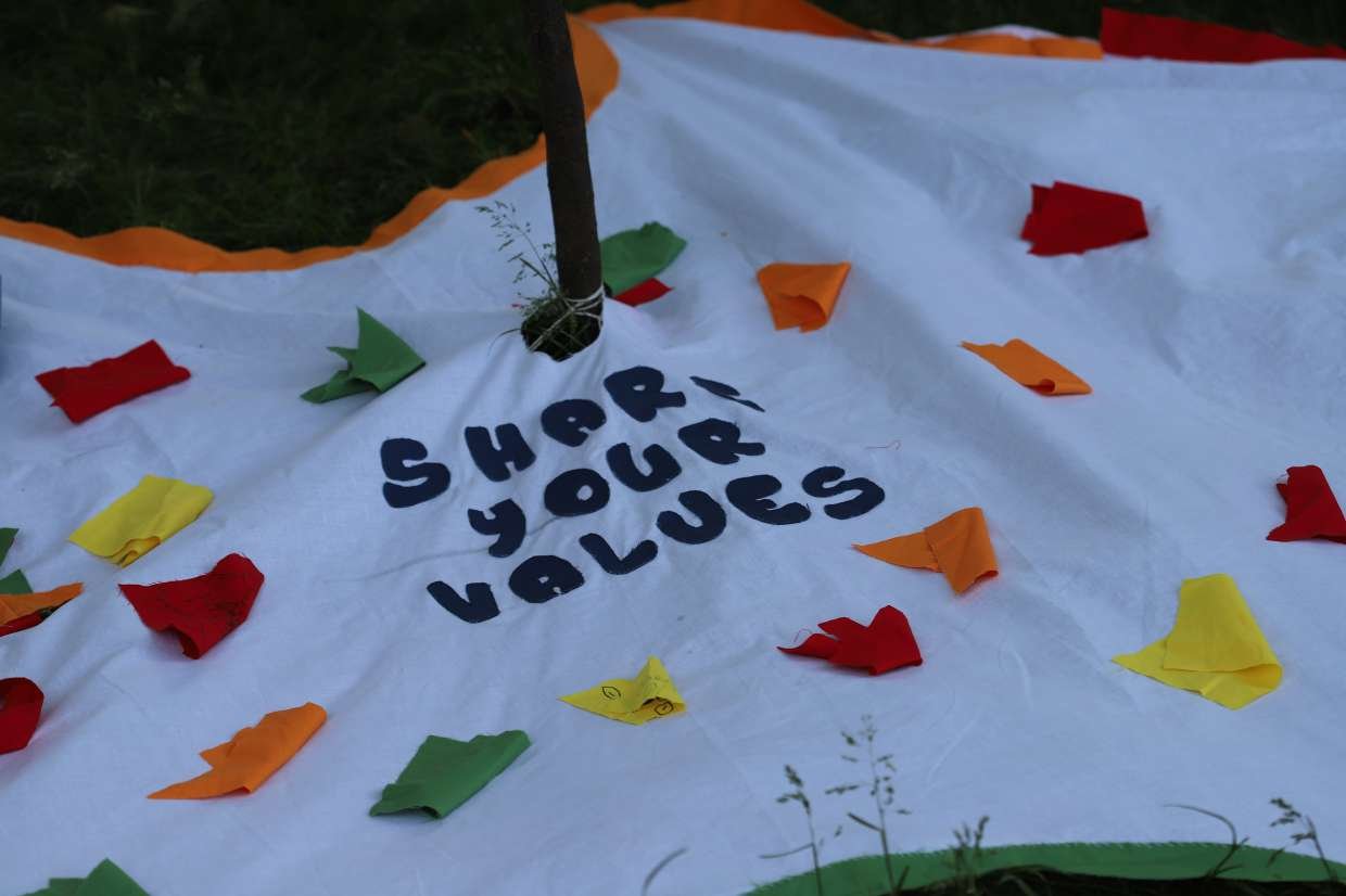 A white fabric surface with the words "SHARE YOUR VALUES" written in dark blue letters, decorated with colorful paper triangles and confetti, and a pole in the center.