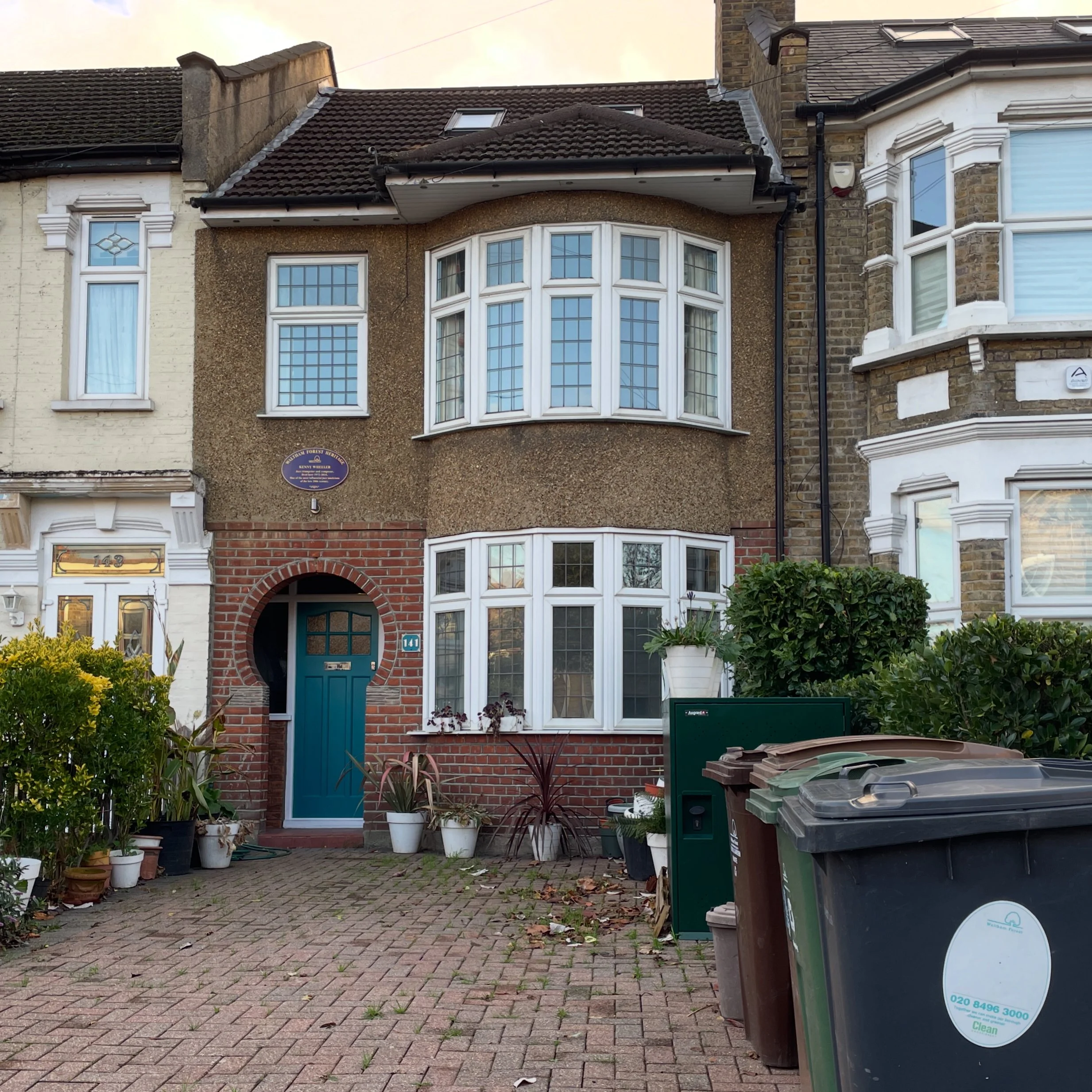 A three-story house with a brick and gravel exterior, blue front door, bay windows, and potted plants in front. There are trash bins and bushes on the driveway.
