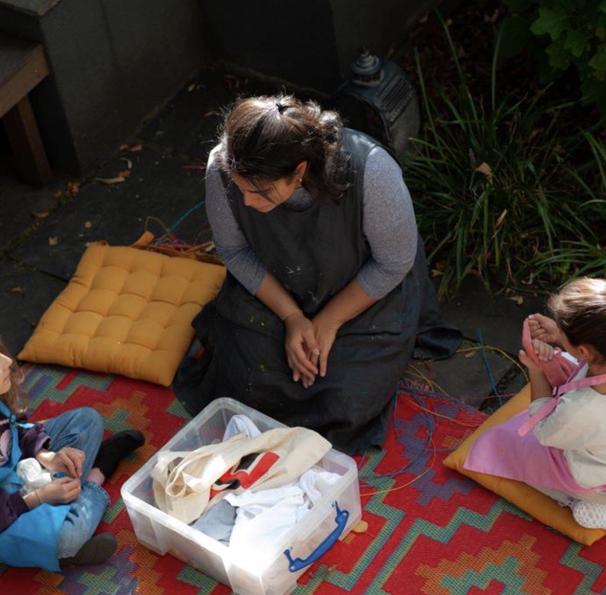 Woman sitting on a colorful rug with children, surrounded by cushions and a plastic container filled with cloth and bags, outdoors at night.