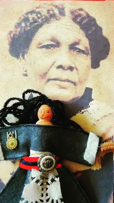 A black and white portrait of an older woman with styled hair in the background, and in the foreground, a doll with black yarn hair, wearing a navy uniform with a badge and medals, sitting in front of the portrait.