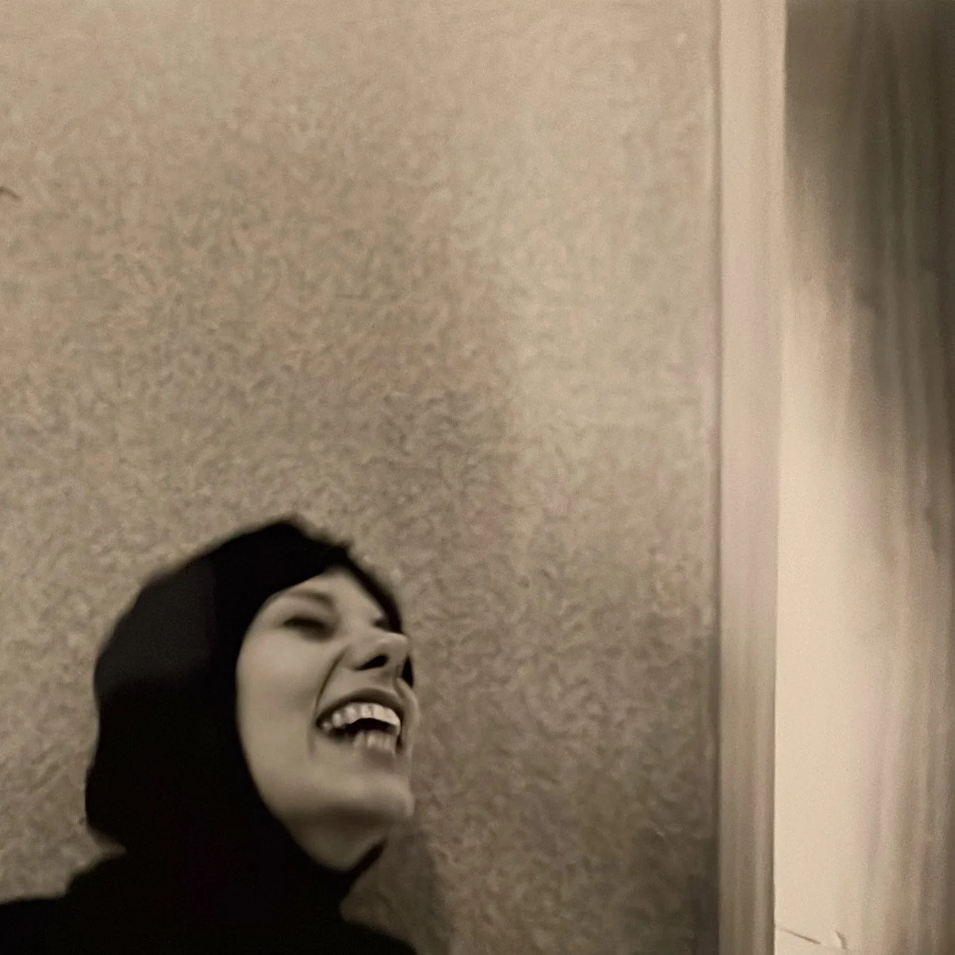 A person with light skin and dark hair, wearing a black hoodie, is laughing and looking up, standing against a textured wall.
