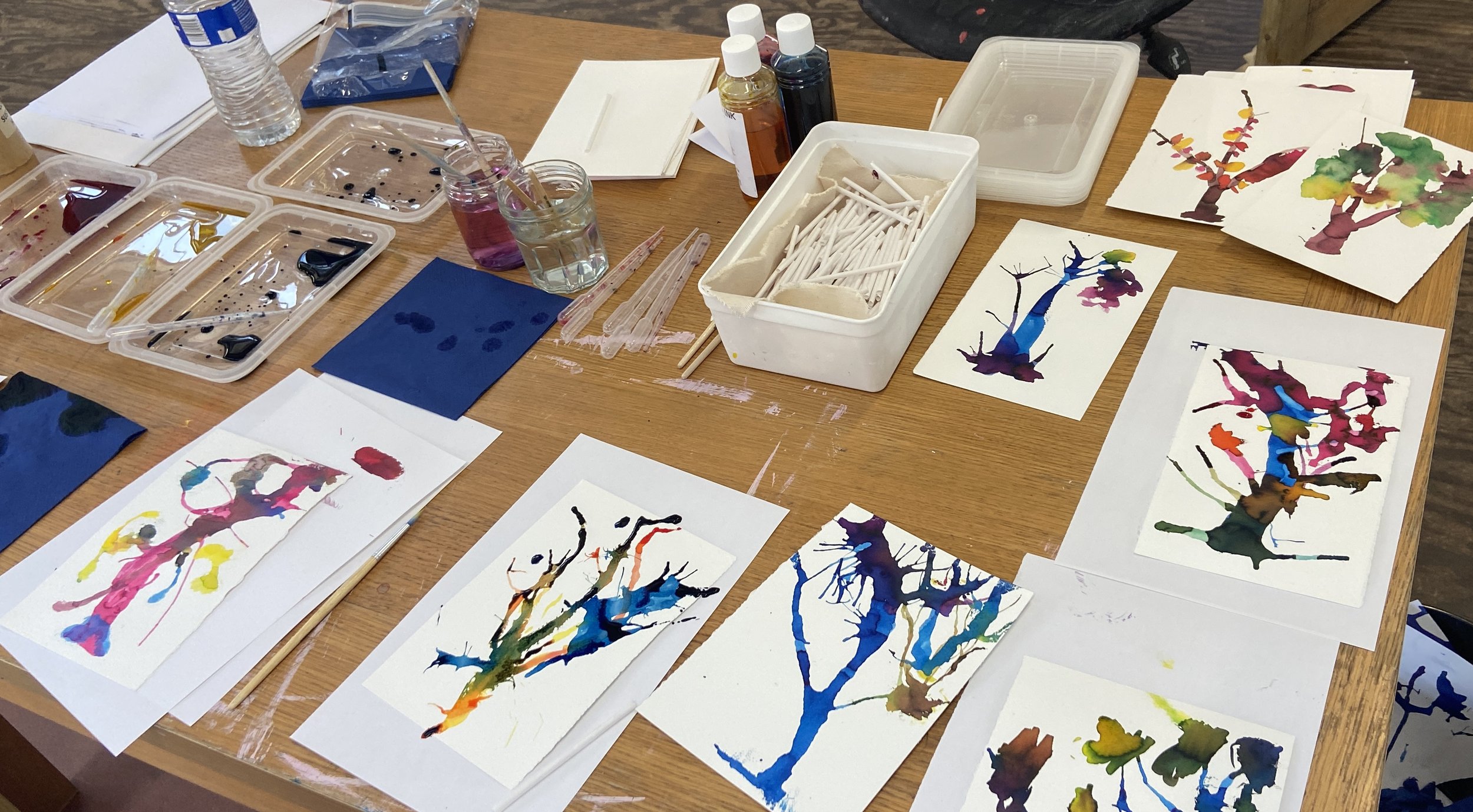 Watercolor paintings of trees on paper, with art supplies like paint, brushes, and water bottles on a wooden table.