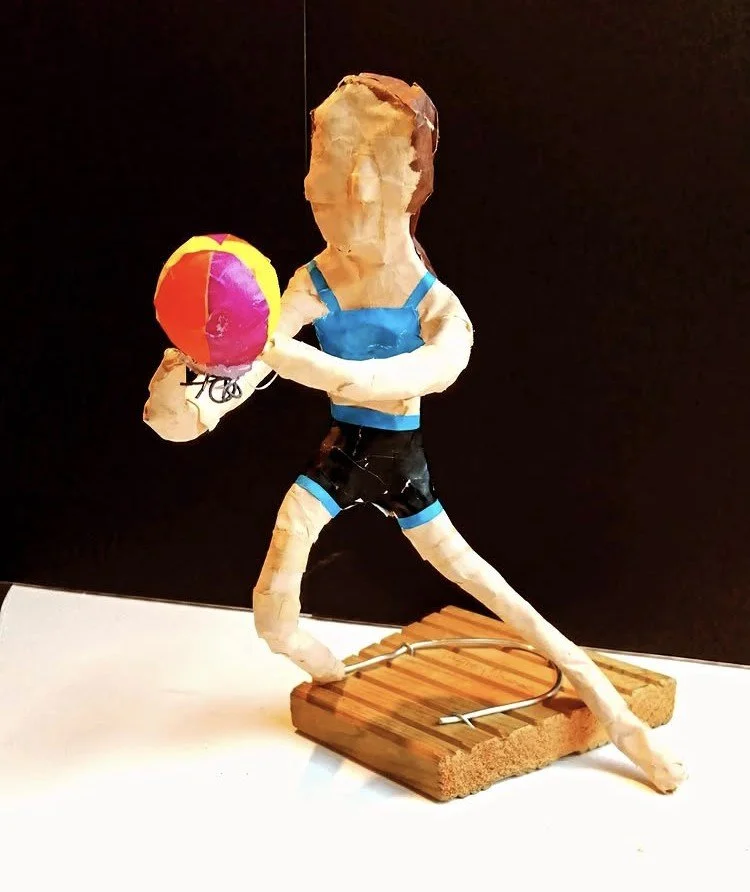 Paper mache sculpture of a person in blue top and black shorts holding a colorful beach ball, standing on a wooden base with a wire structure, against a black background.