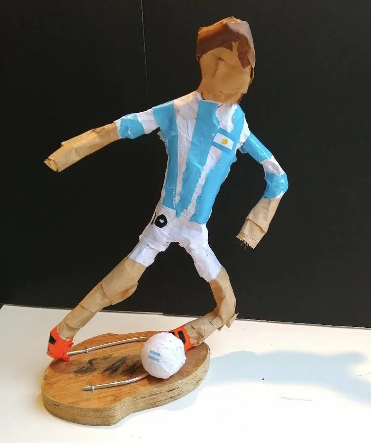 A handcrafted paper sculpture of a soccer player wearing a blue and white striped Argentina jersey with a number and the Argentinian flag, white shorts, orange shoes, and a white soccer ball on a wooden base.