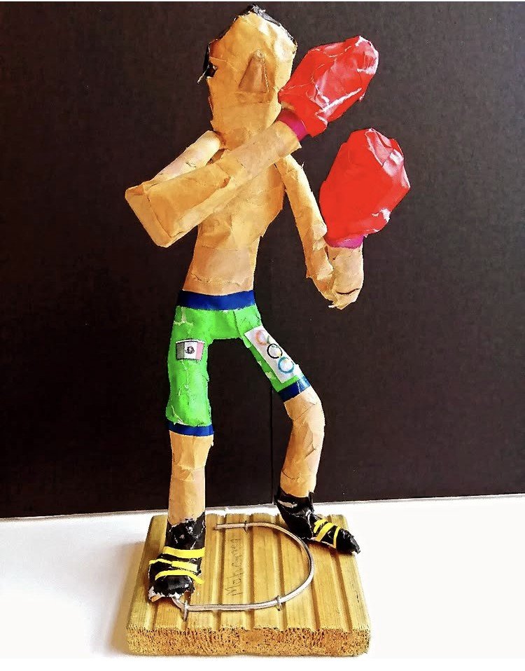 A sculpture of a boxer with a muscular build, standing on a wooden base with a metal ring at his feet. The figure is wearing green shorts with blue trim and boxing gloves, with a black and yellow boxing shoes. The sculpture is made from crumpled paper and tape, with a black background.