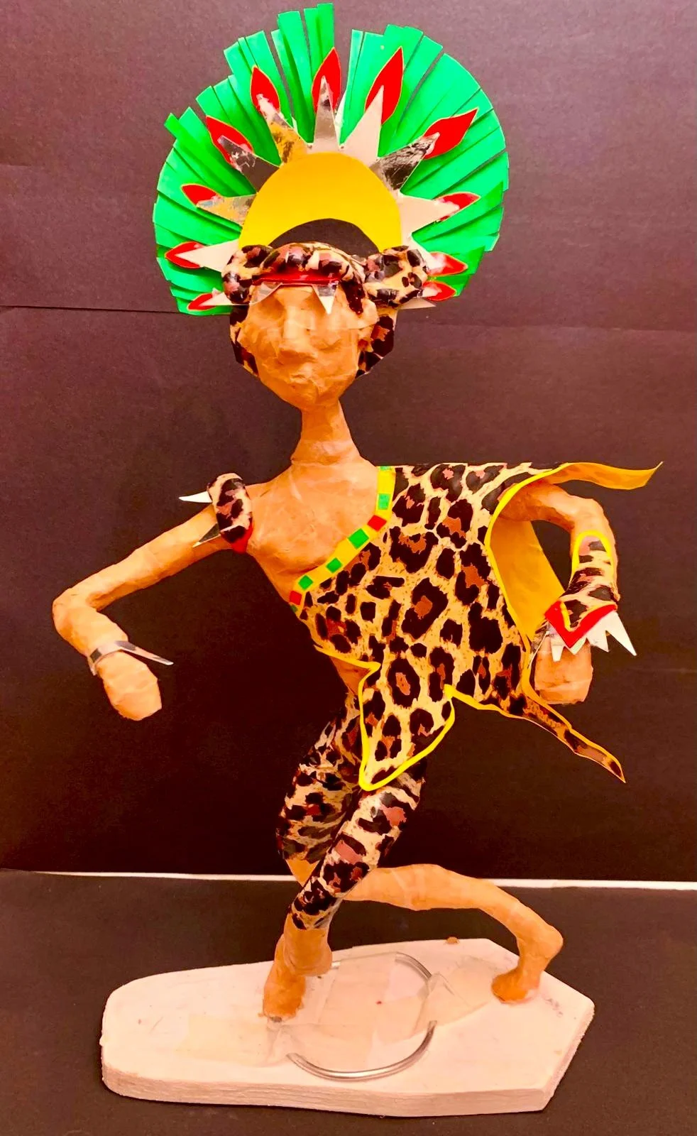 A colorful sculpture of a person dressed in animal print clothing, wearing a vibrant headdress resembling a sun with rays, standing on a white base against a dark background.