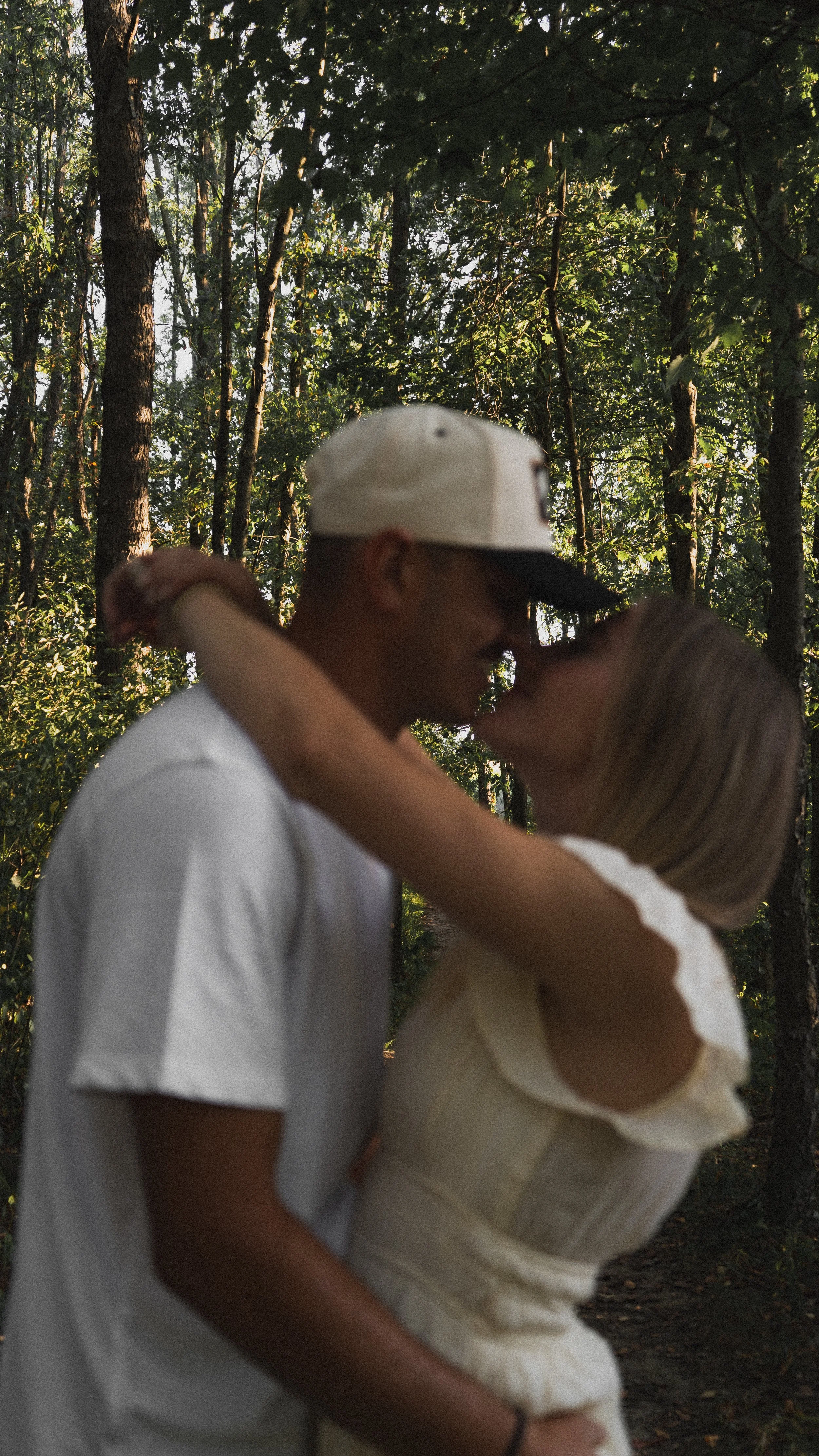 A young couple kissing in a forest, with trees and sunlight filtering through the leaves in the background.