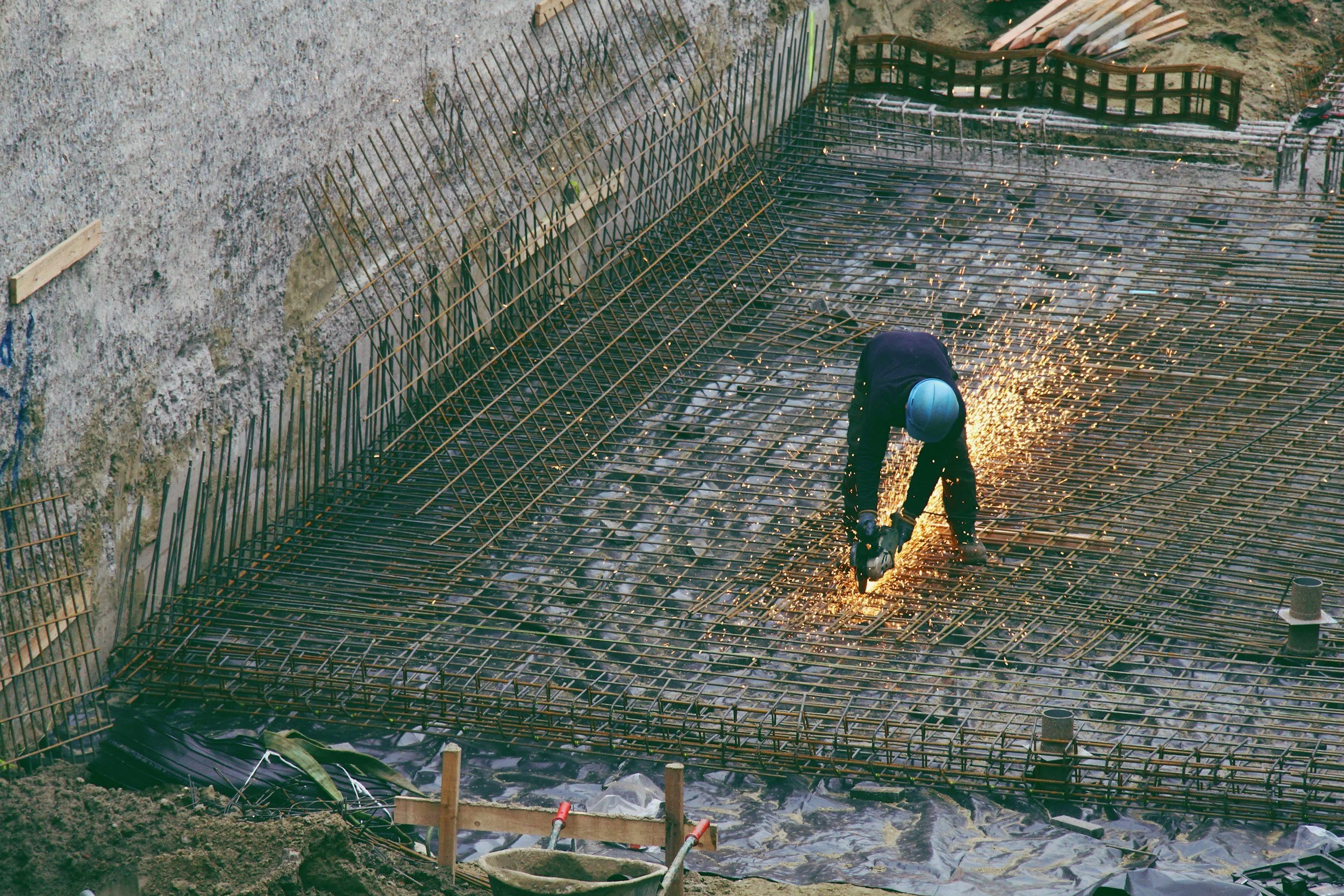 Construction worker welding rebar on a building foundation, sparks flying, wearing a blue helmet and dark work clothes.