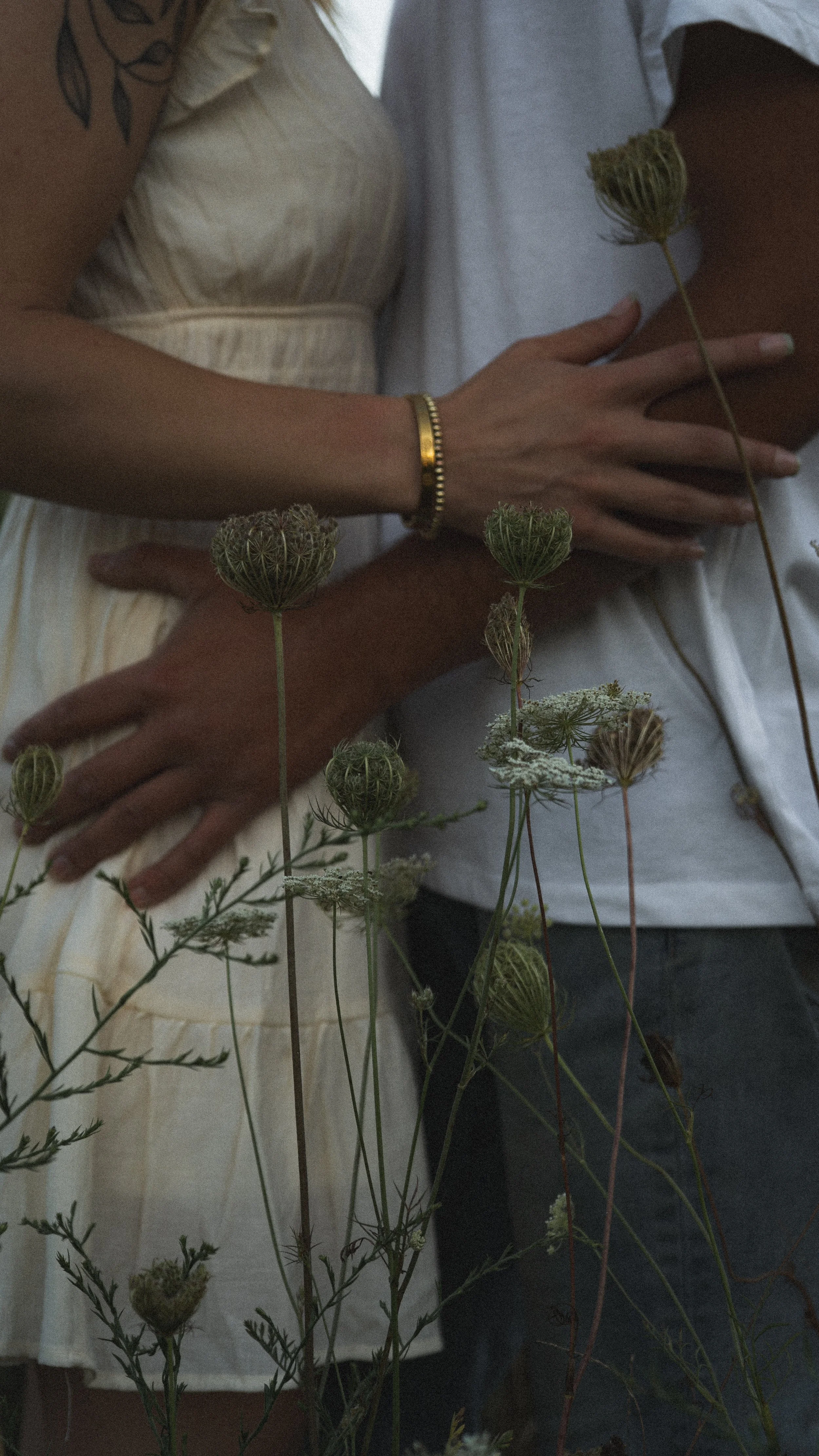 A couple embracing outdoors with wildflowers in the foreground, the woman wearing a light-colored dress and a gold bracelet, the man in a white shirt.