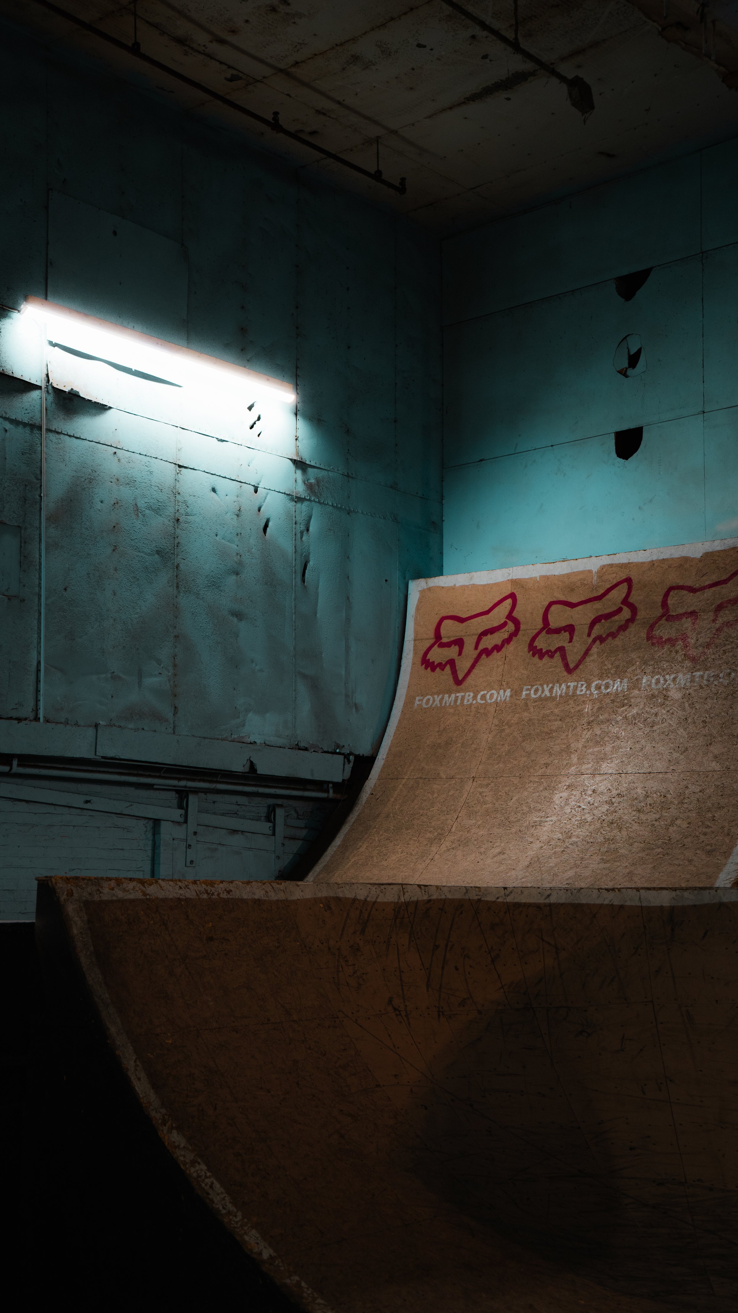 Indoor skatepark with a half-pipe ramp featuring Fox Racing logos, dim lighting, and industrial-style walls.