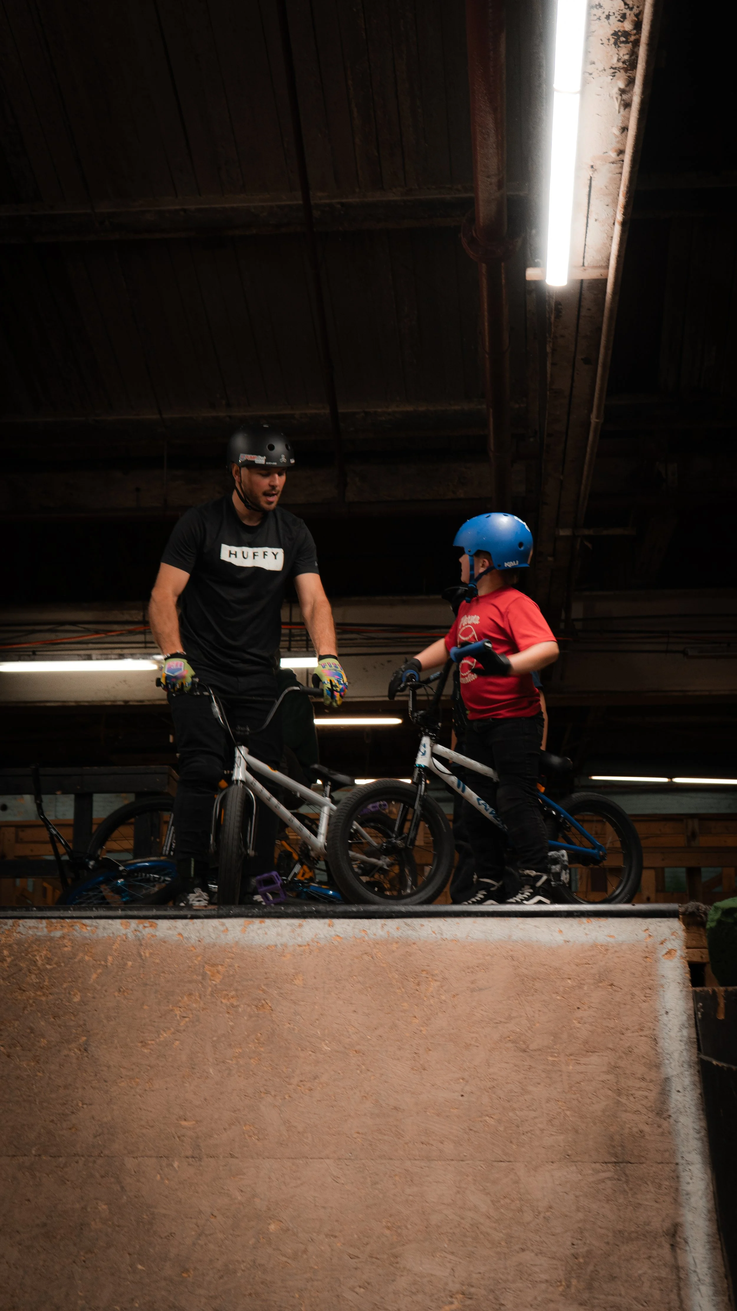 An adult man and a young boy, both wearing helmets, standing with their bikes inside an indoor skate park or BMX riding area, engaging in conversation.