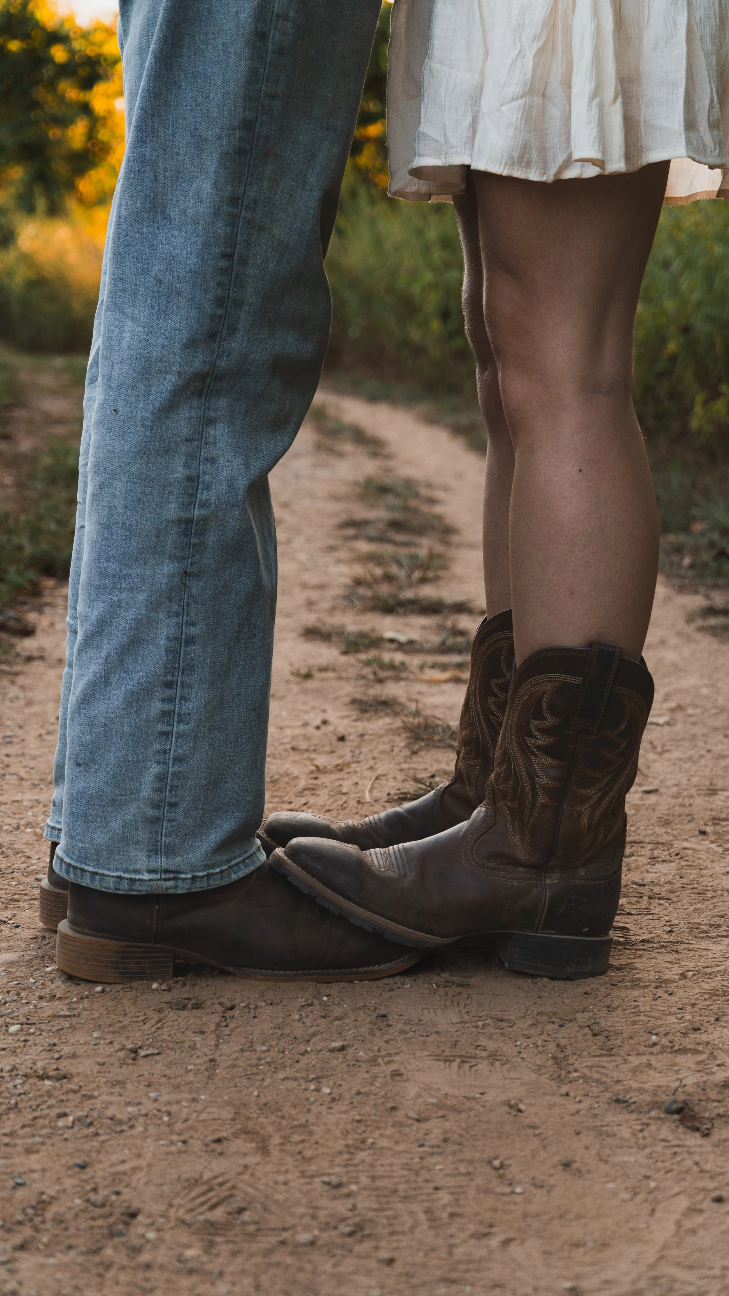 Two people standing close together, the person on the left wearing blue jeans and black shoes, the person on the right wearing a white dress and cowboy boots on a dirt path surrounded by greenery.