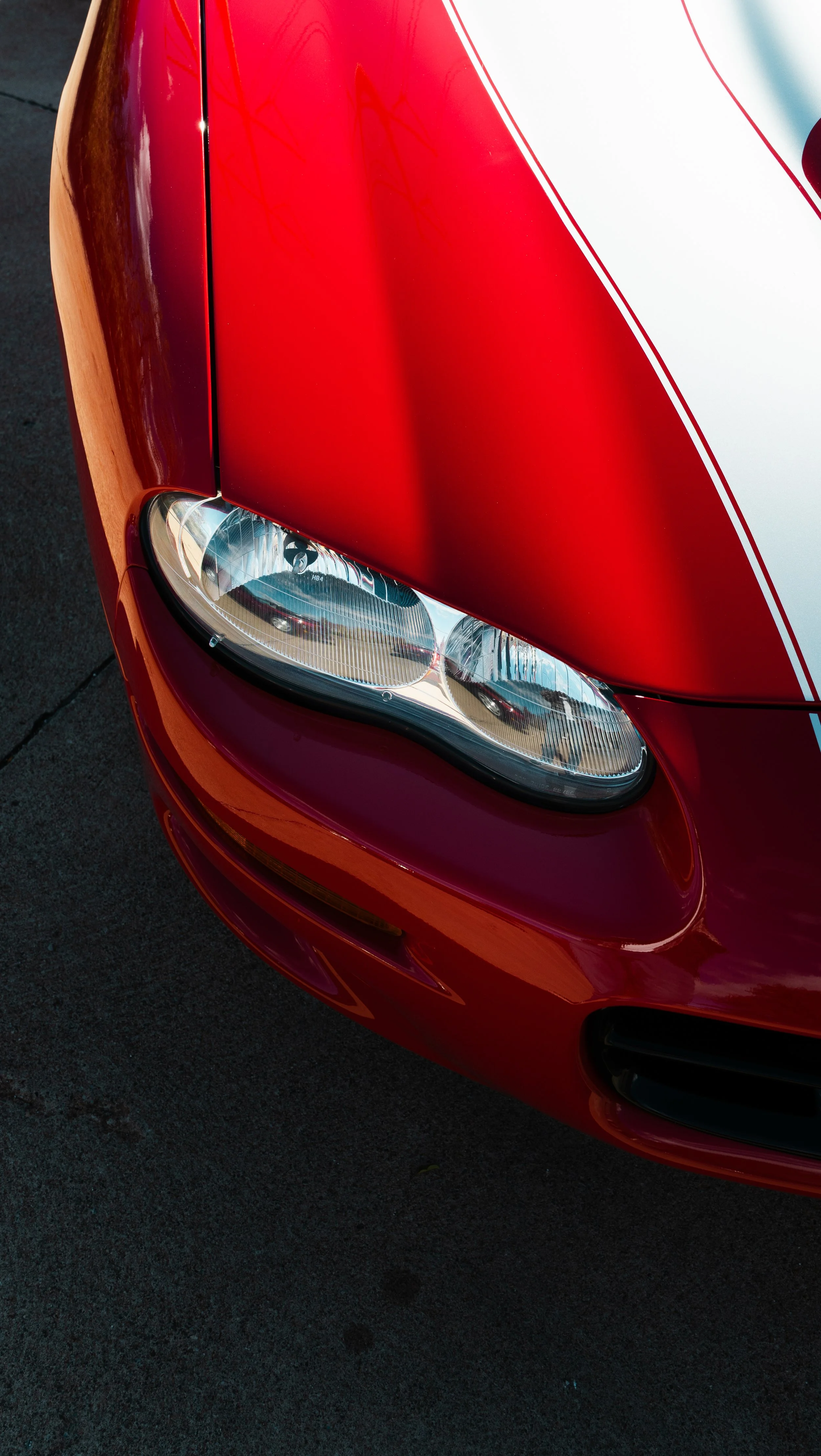 Close-up of the front left corner of a red sports car with a distinctive headlight and white racing stripes.