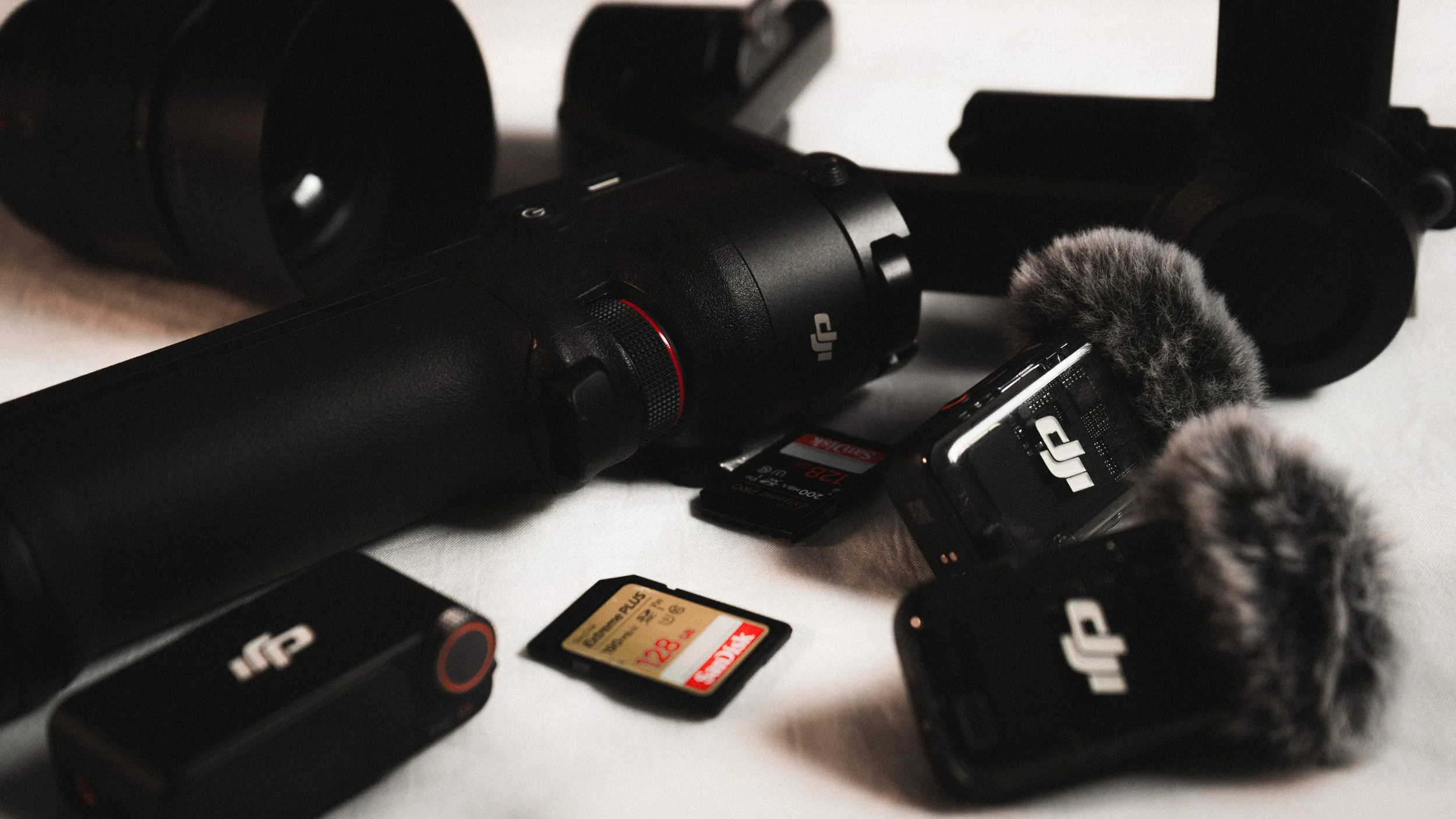 Photography equipment including a large camera stabilizer, two microphones with furry windshields, a memory card, and a small black device, all placed on a white surface.
