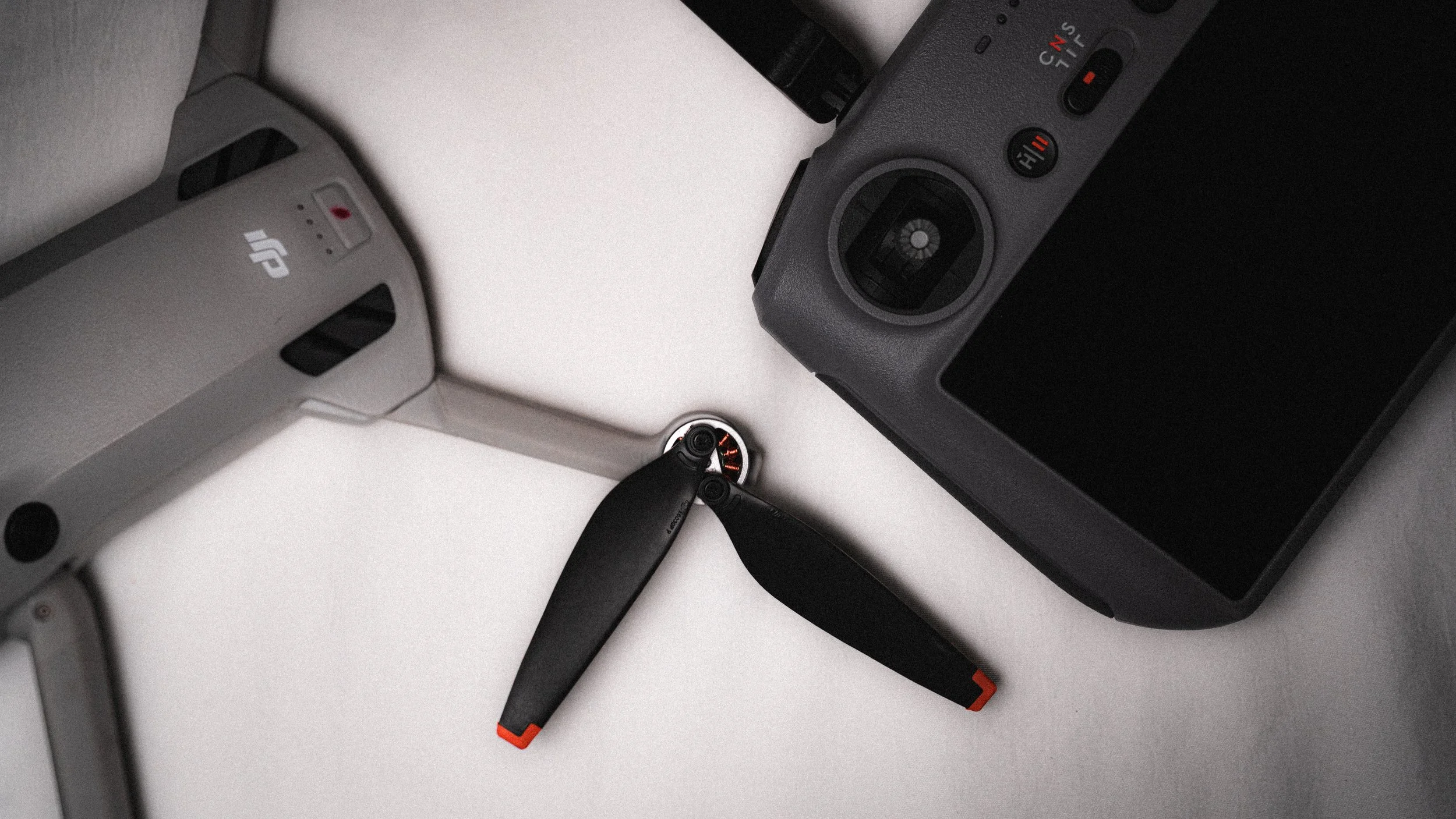 A drone with a black propeller and camera, a DJI drone's remote control, and a black camera are on a white surface.