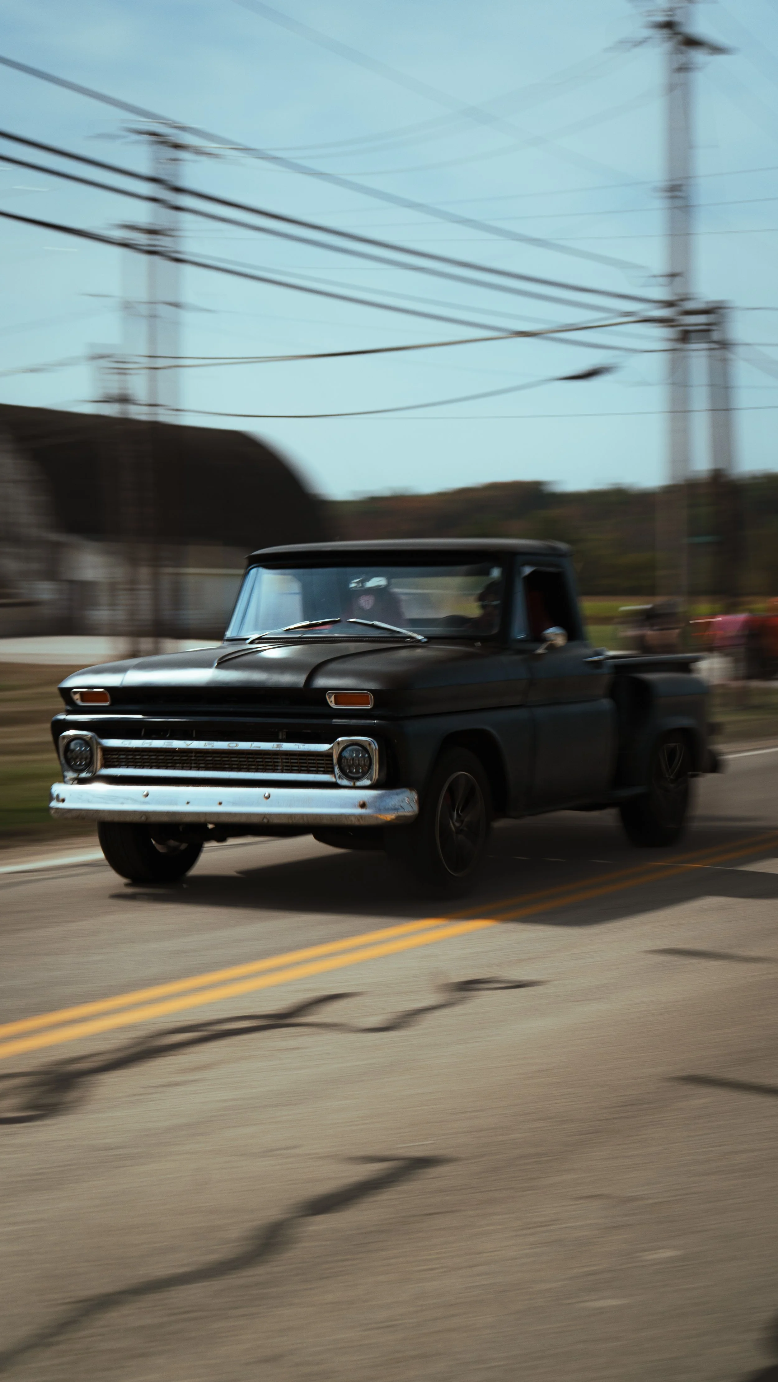 A black vintage pickup truck driving on a suburban street with power lines overhead and blurred background.