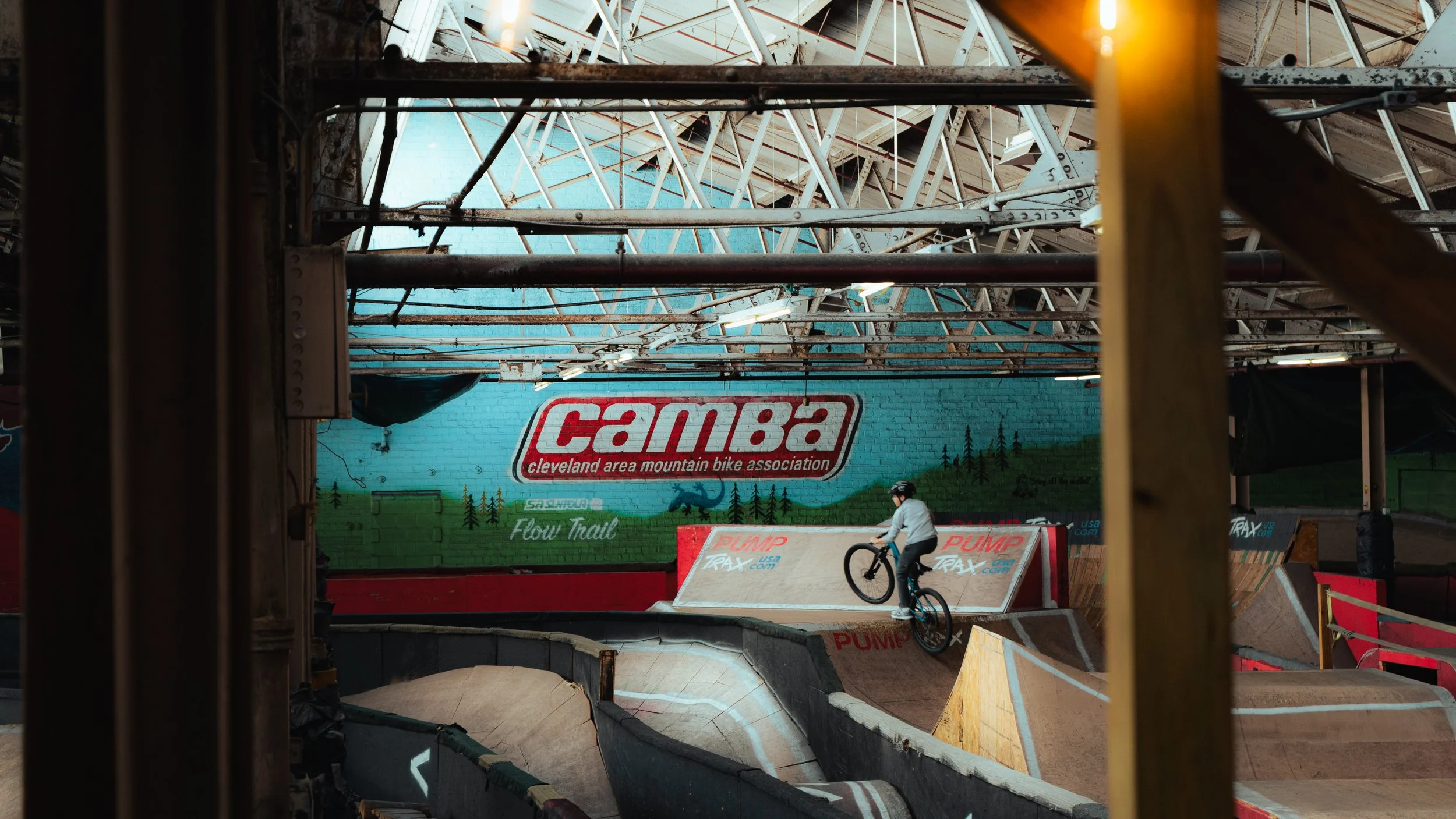 Indoor BMX bike park with a rider performing a jump on his bike on a ramp, surrounded by various ramps and a large Cleveland area mountain bike association banner on the painted brick wall.